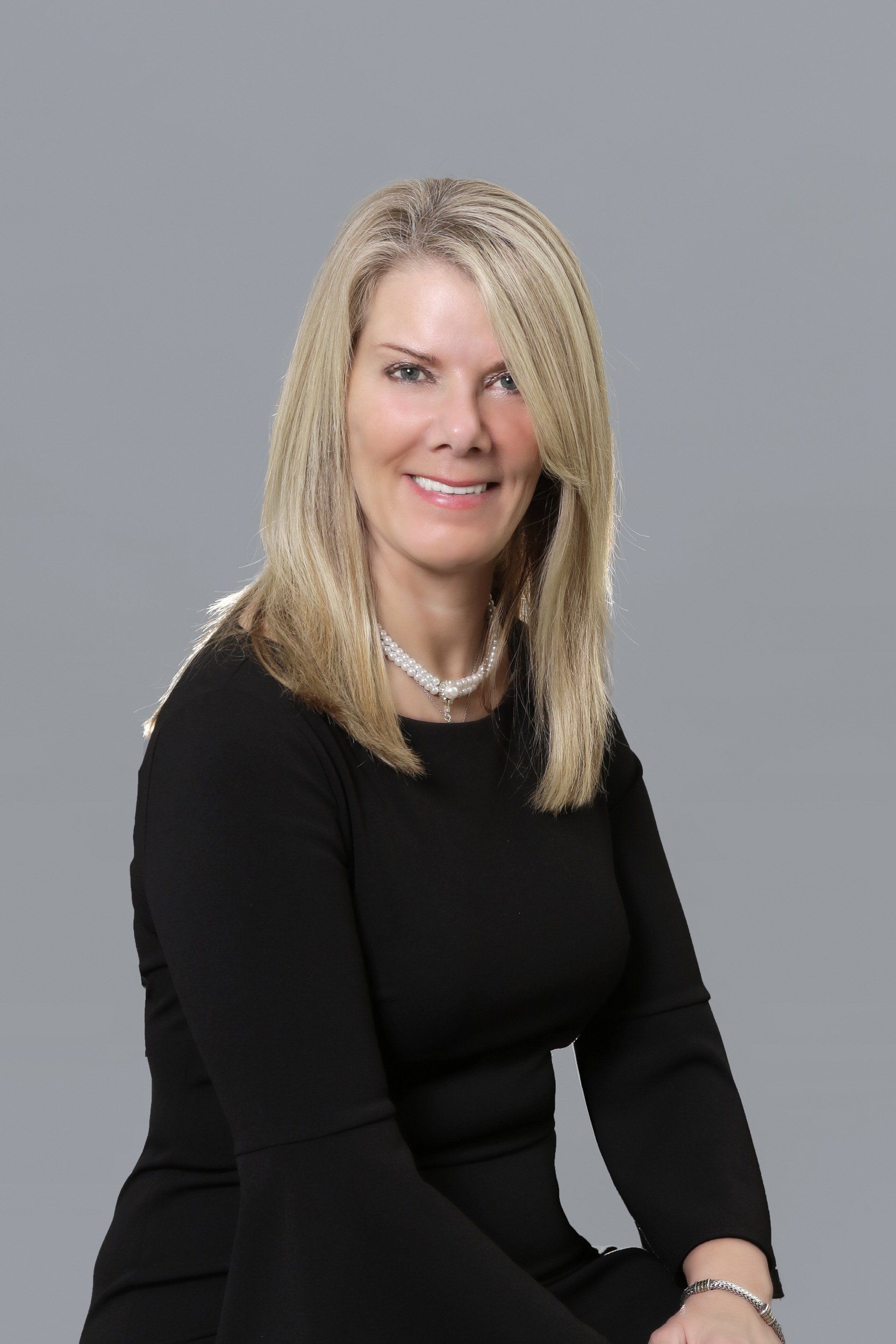 Woman with blonde hair, smiling, wearing a black dress and pearl necklace. Gray background.