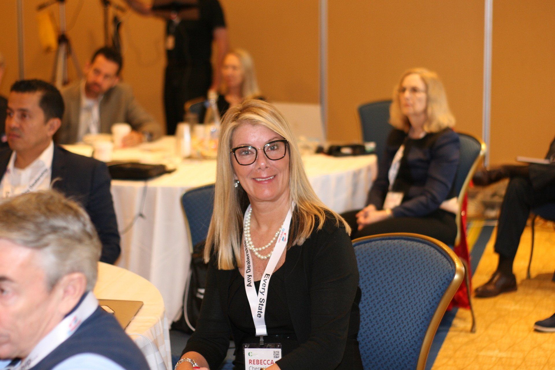 Woman smiling, wearing glasses and a lanyard, seated at a conference, looking at camera.