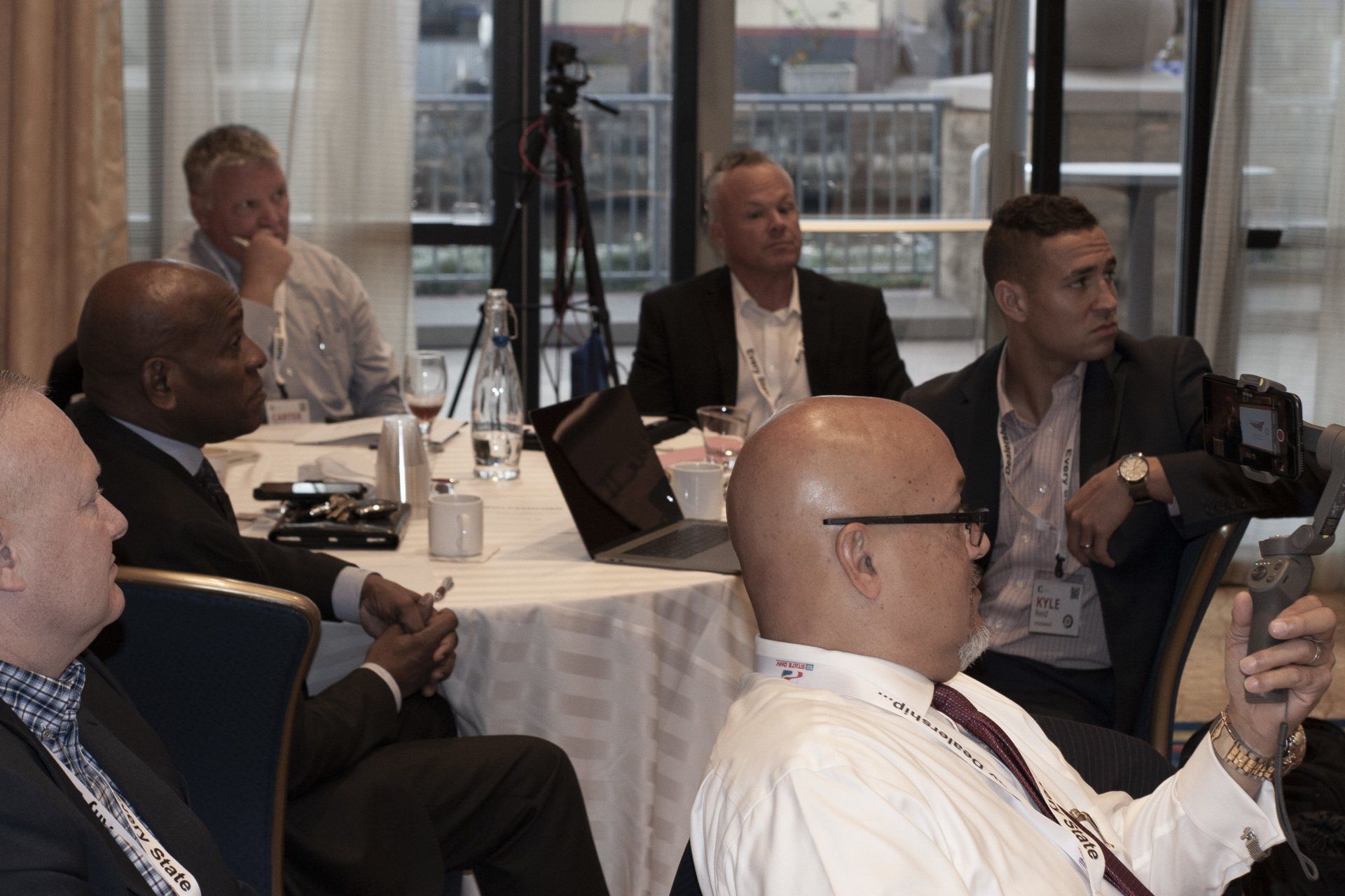 People seated at a round table, listening to a presentation. The setting is likely a conference room or meeting space.