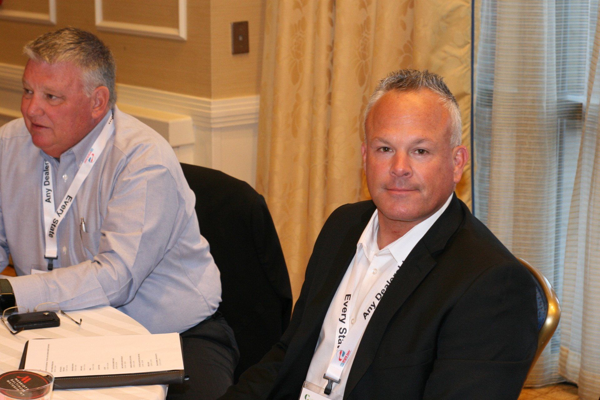 Two men seated at a table. One in a light blue shirt, the other in a black blazer and white shirt, both wearing lanyards.