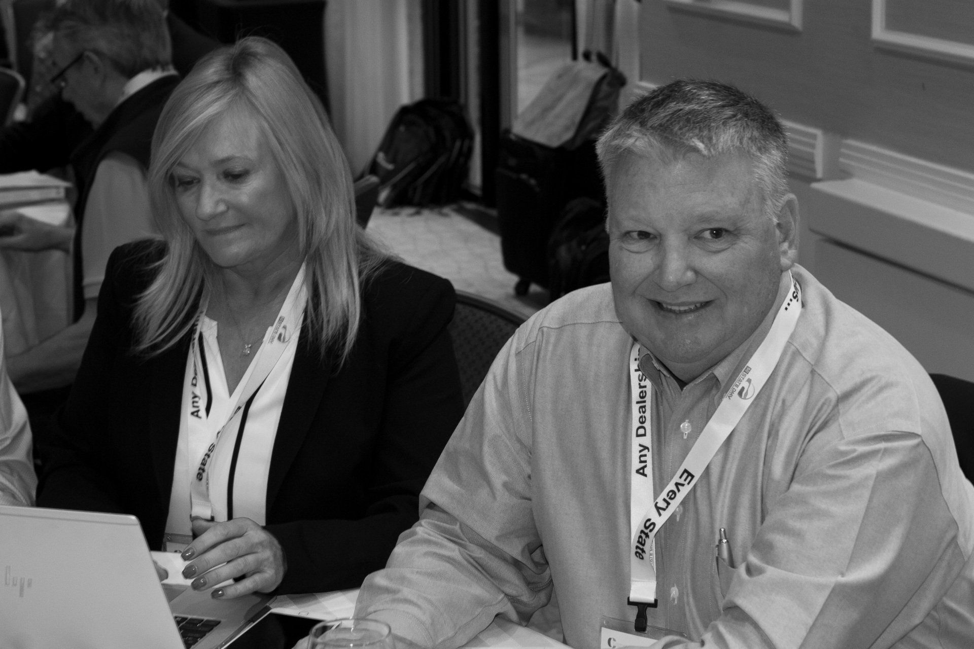 Two people with lanyards sit at a table. The man smiles. Woman looks down.