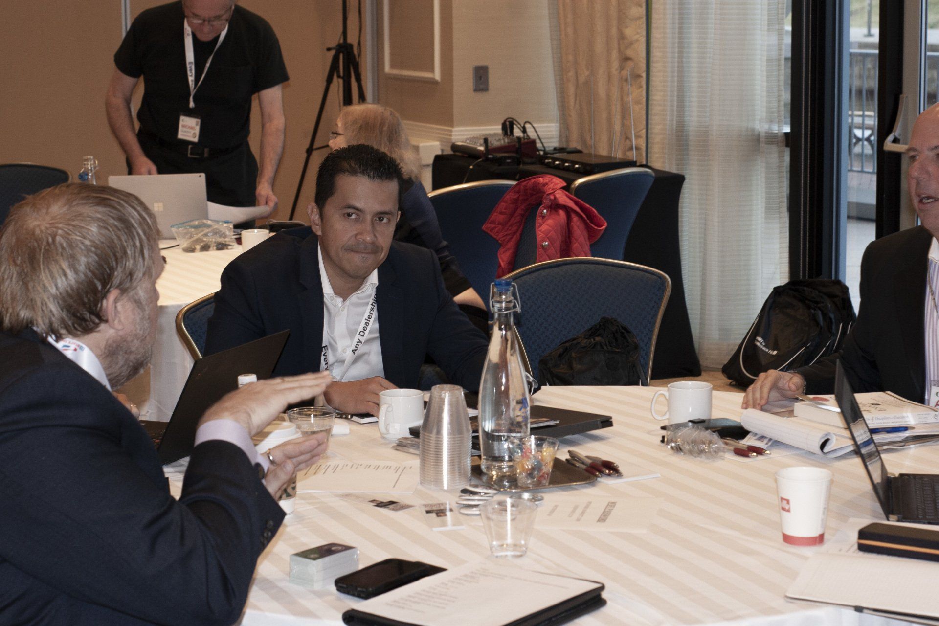 Group of people at a round table, possibly a meeting, with laptops and cups.