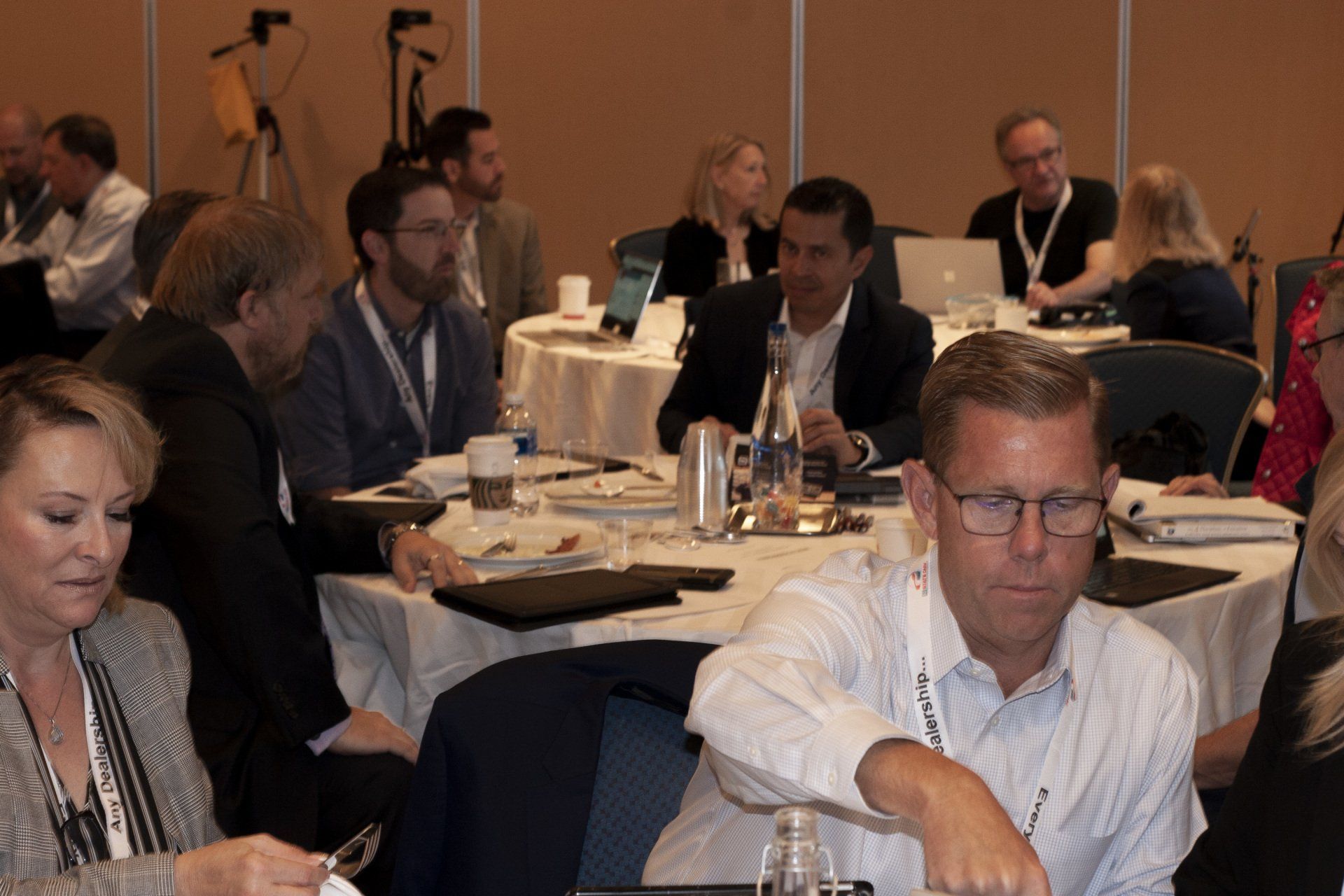 People seated at round tables in a conference setting, some using laptops and taking notes.