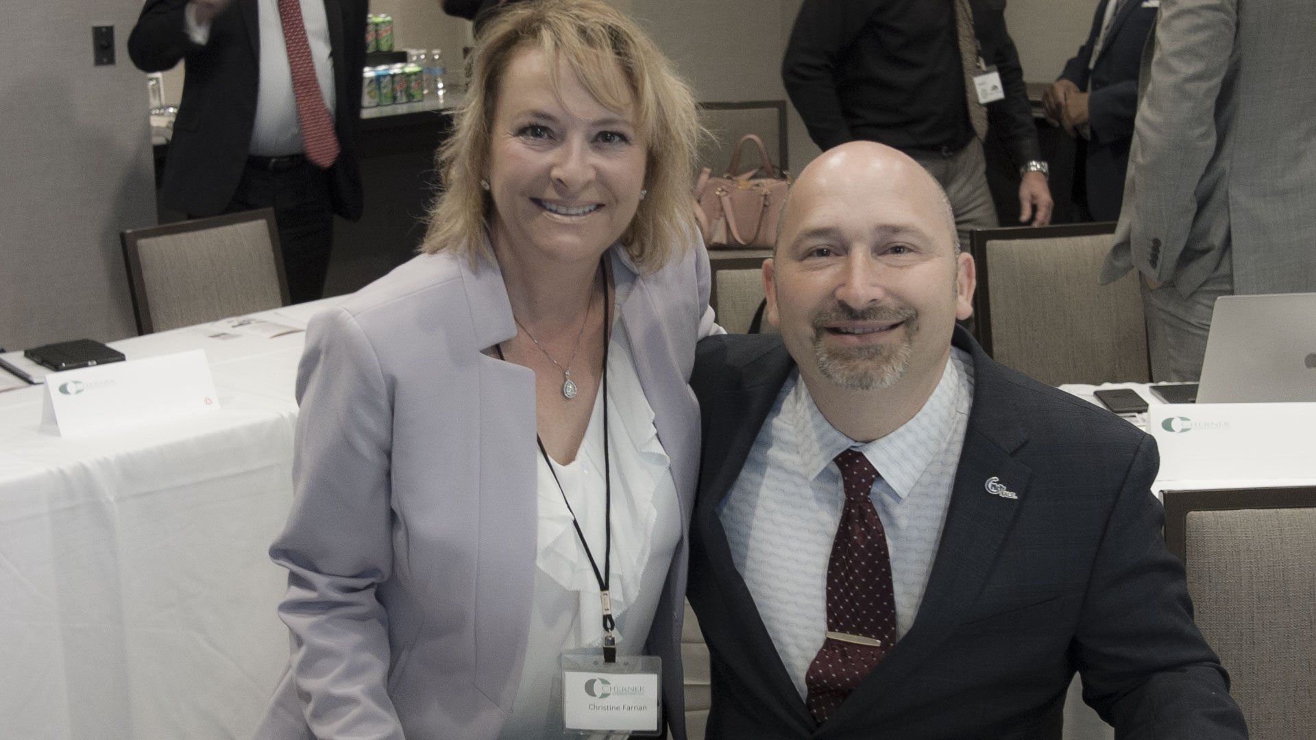 A woman and a man smiling together in a room with a white table. The man has a tie.