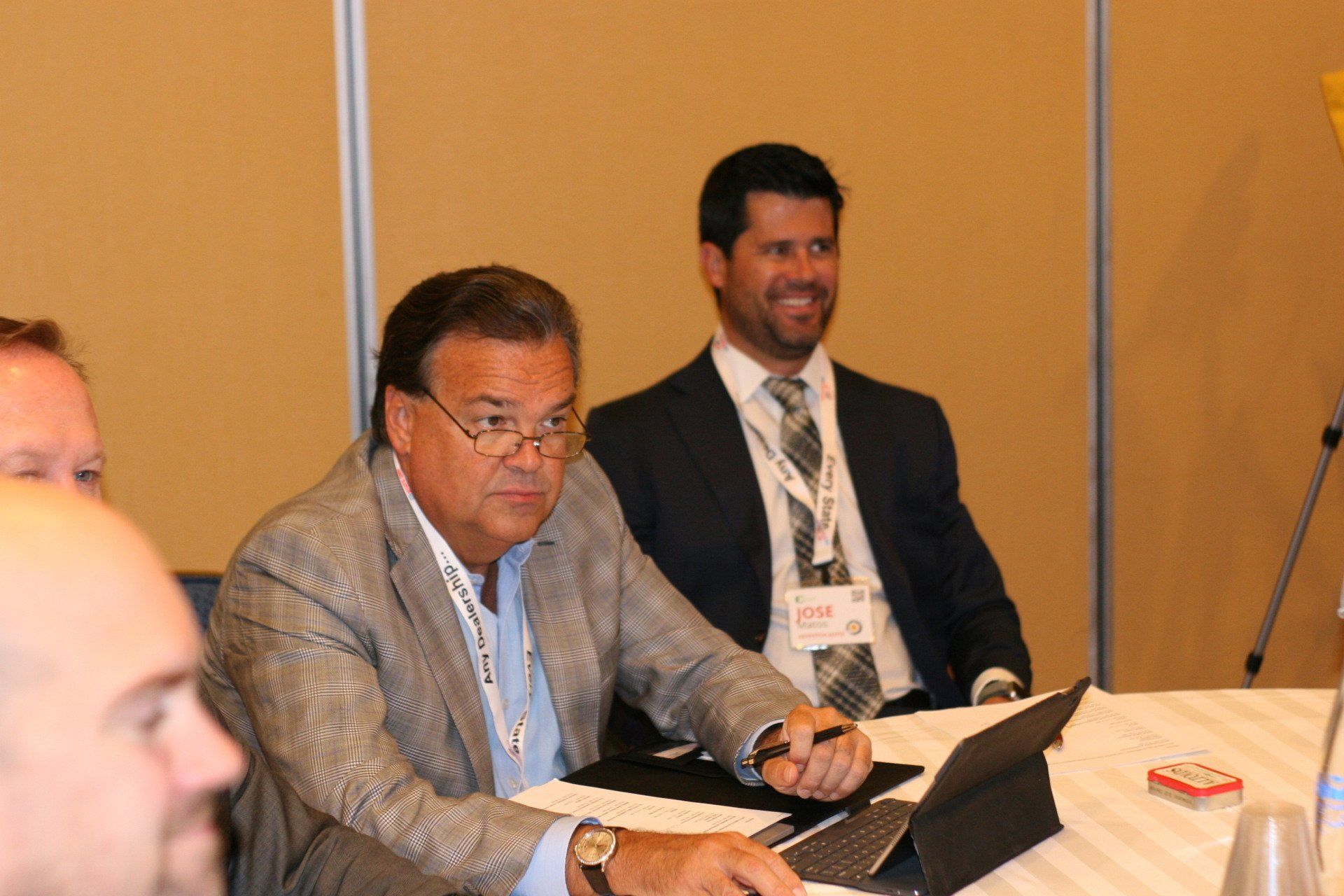 Two men at a conference table; one looks at a tablet, the other smiles. Beige wall background.