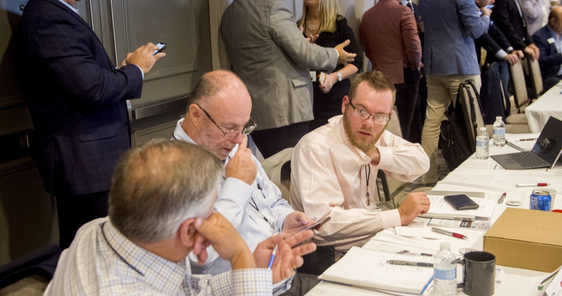 Attendees at a conference table, looking at papers and devices. Others stand nearby.