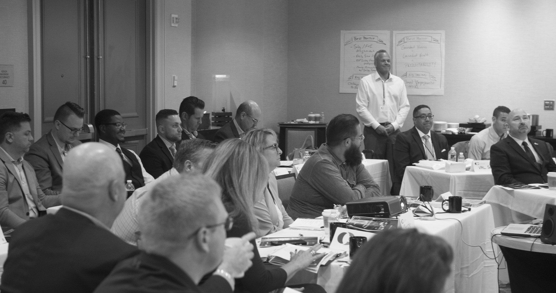Group of people in a meeting room, listening to a speaker standing near a whiteboard.