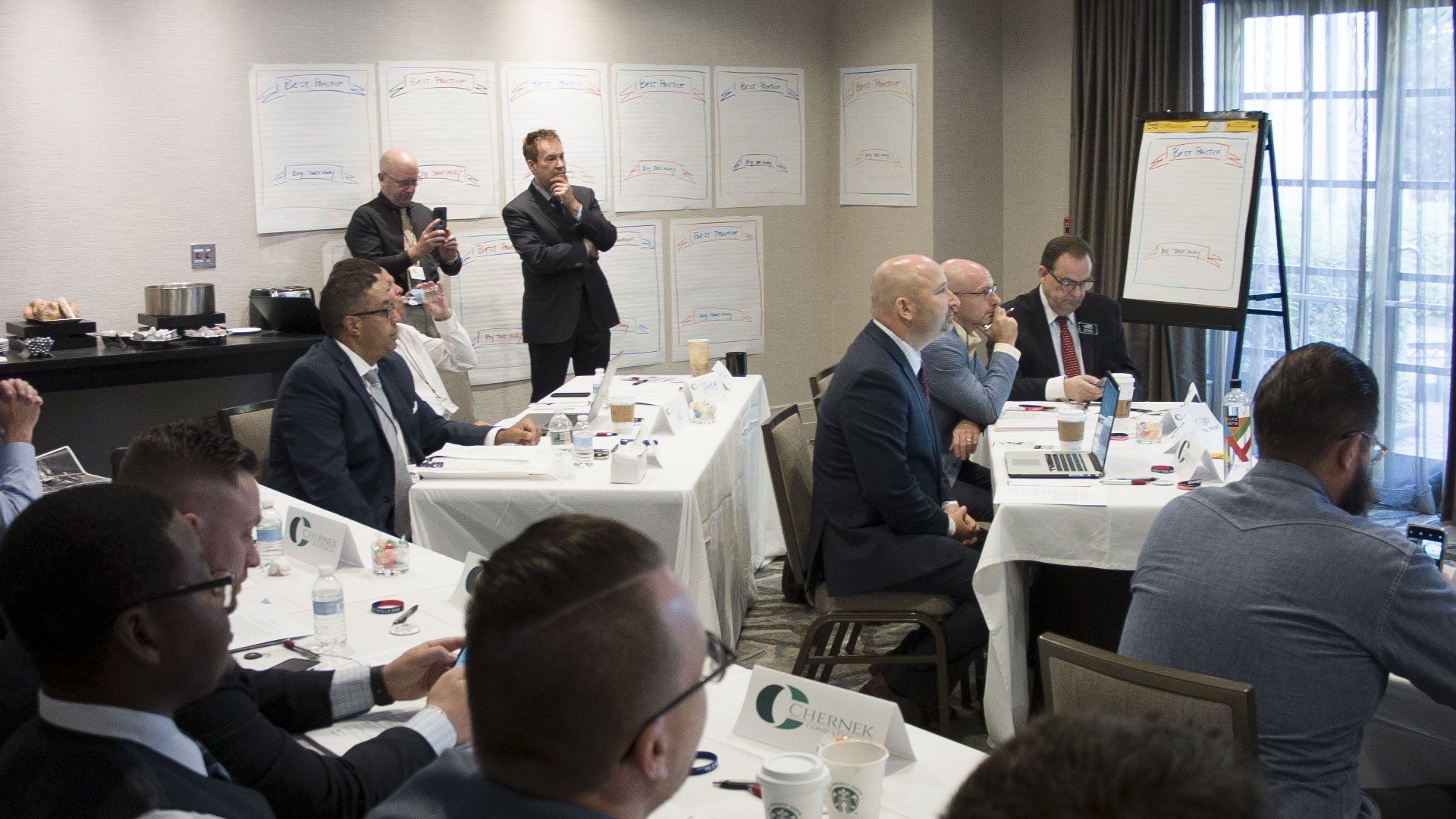 Business meeting in a conference room. People seated at tables, one man stands with hands clasped. Whiteboards in background.