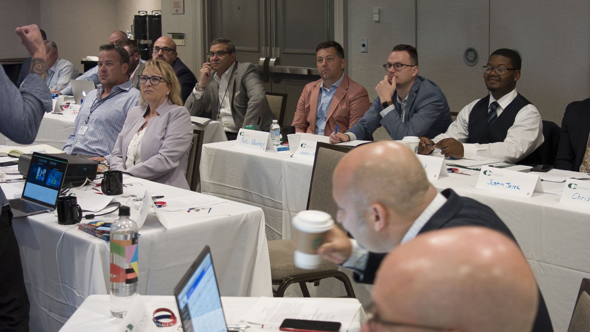 People seated at a conference table, listening. One person is gesturing. Several laptops and water bottles on the table.