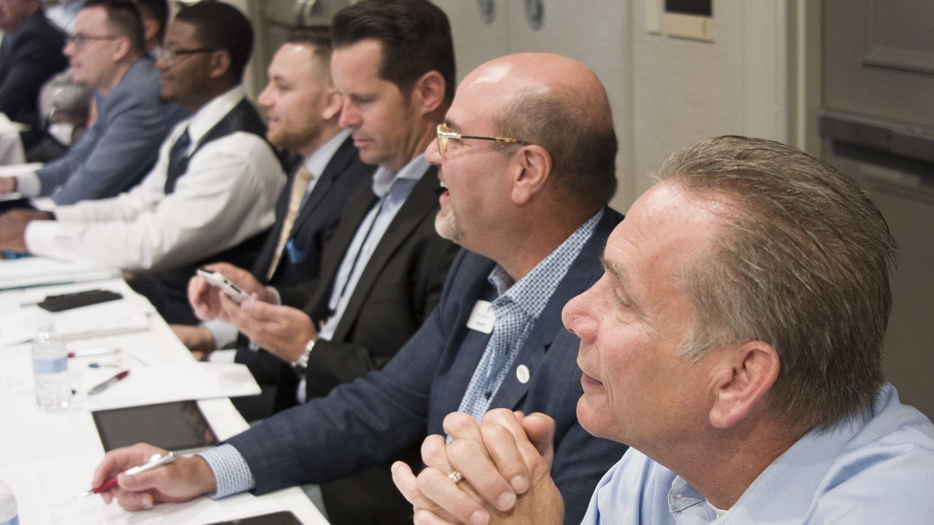 Men in suits seated at a table, some looking forward, others holding phones, in a conference room.