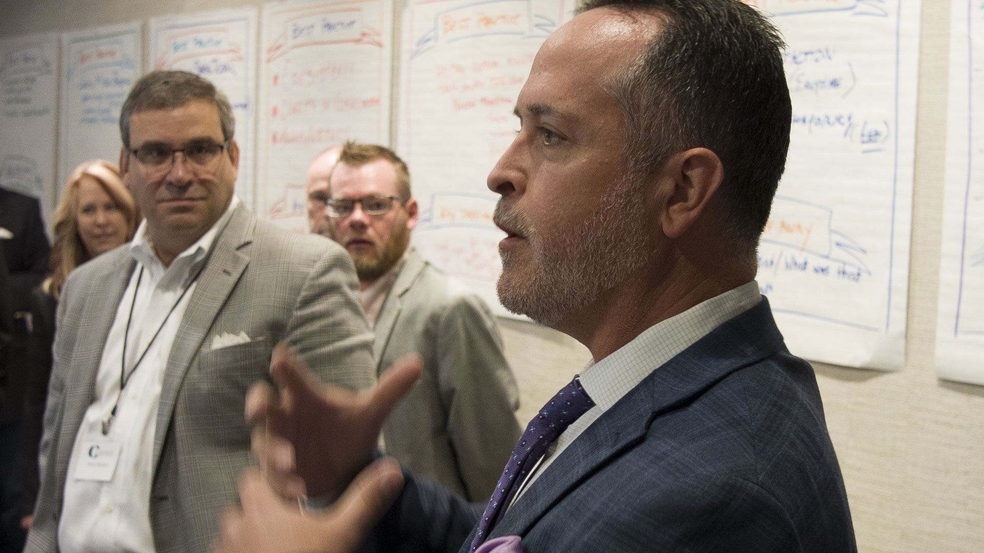 Man in blue blazer gesturing, speaking to group near whiteboard.