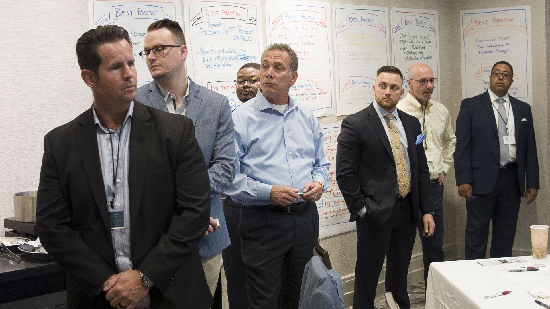 Group of men in business attire listening in a conference room with whiteboard notes.