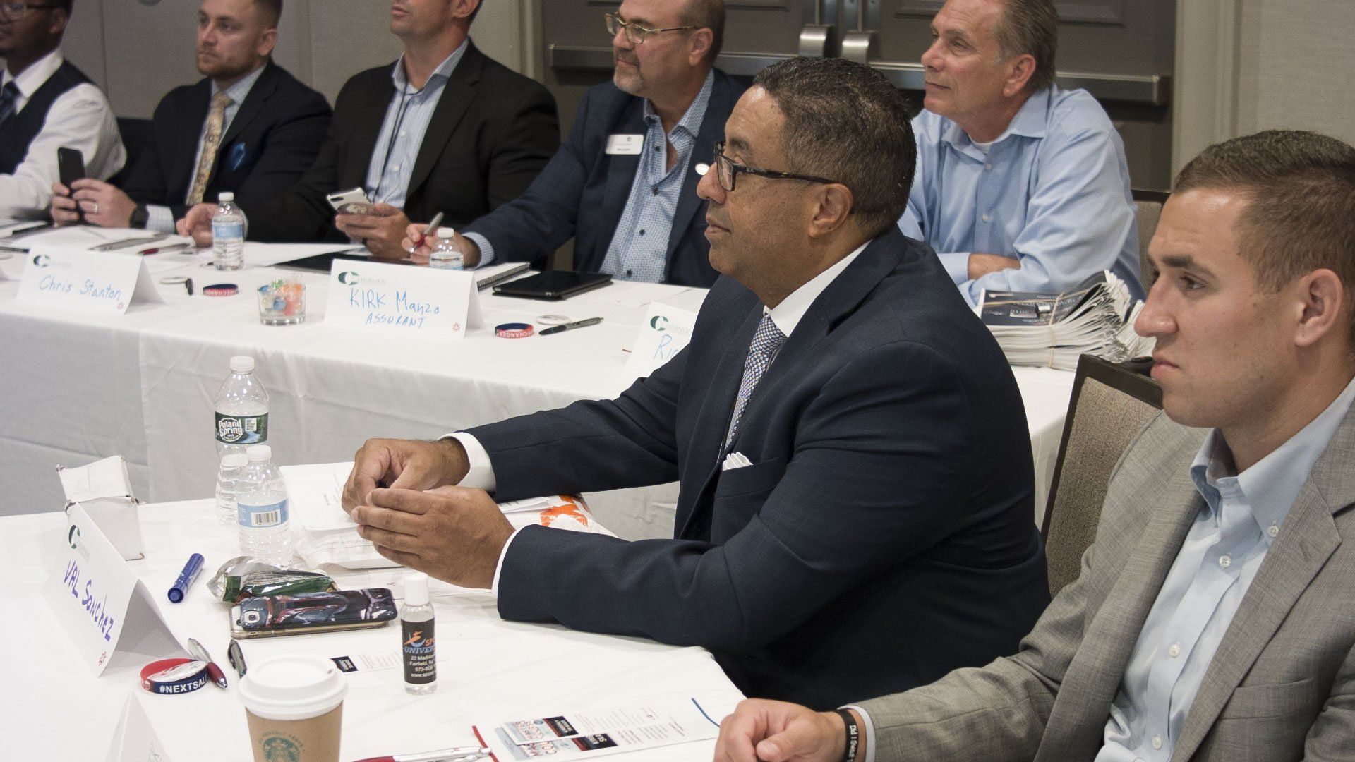 People seated at a table at an event, some wearing suits, listening attentively.