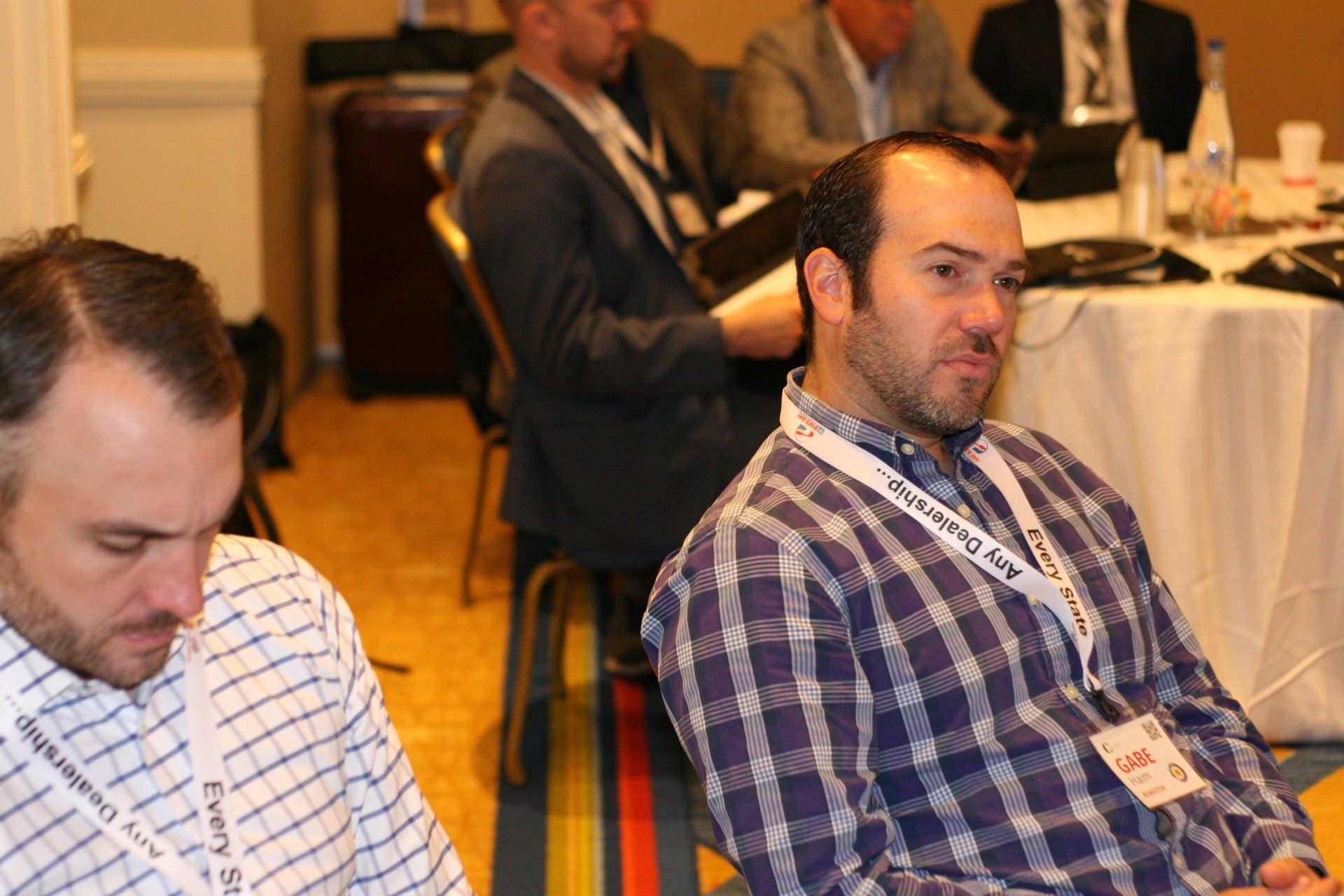 Men in a conference room; one in plaid shirt looks intently ahead, the other looks down. Others sit behind a table.