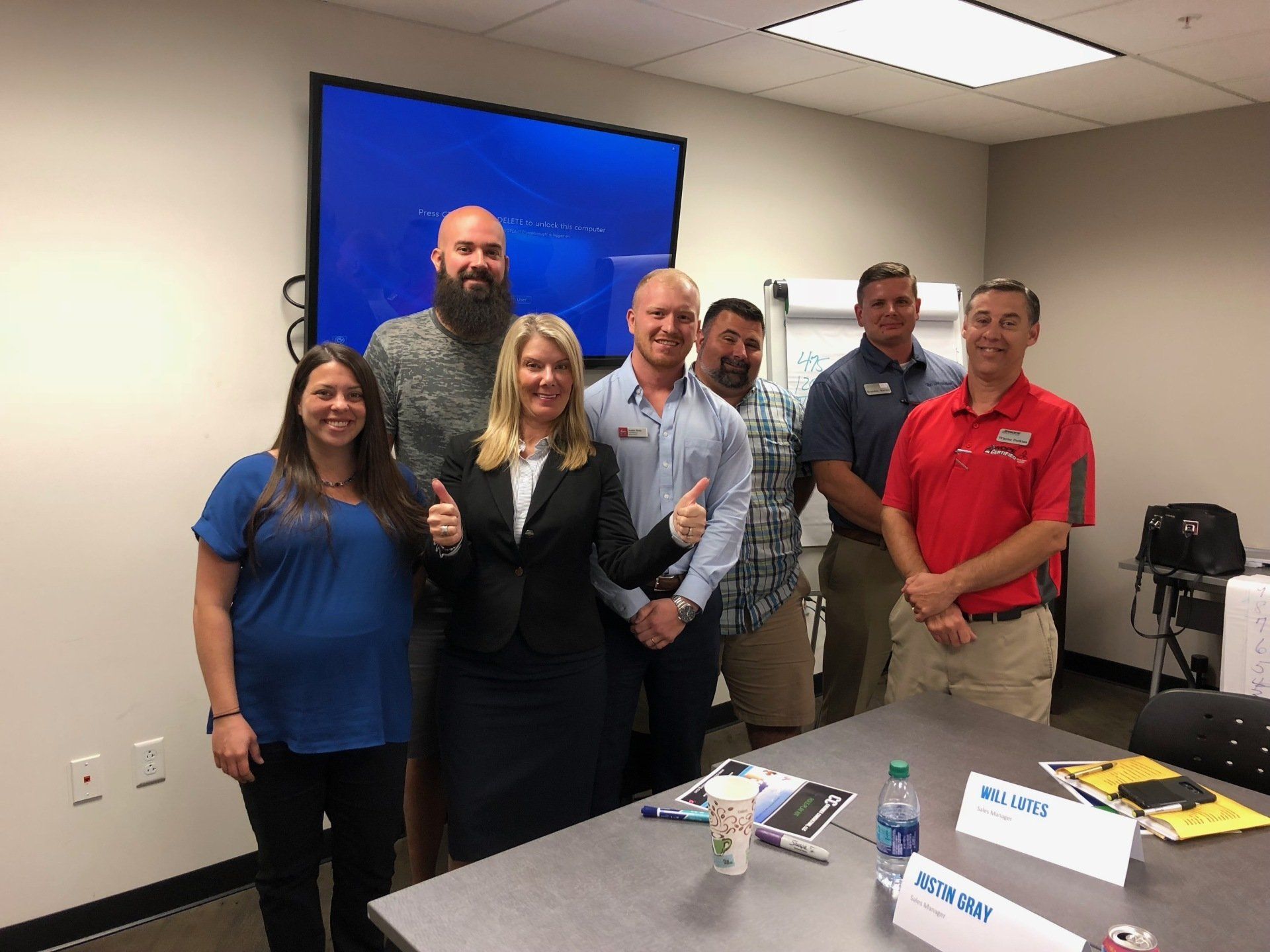 Group of people in a meeting room, some giving thumbs up. A screen is behind them.