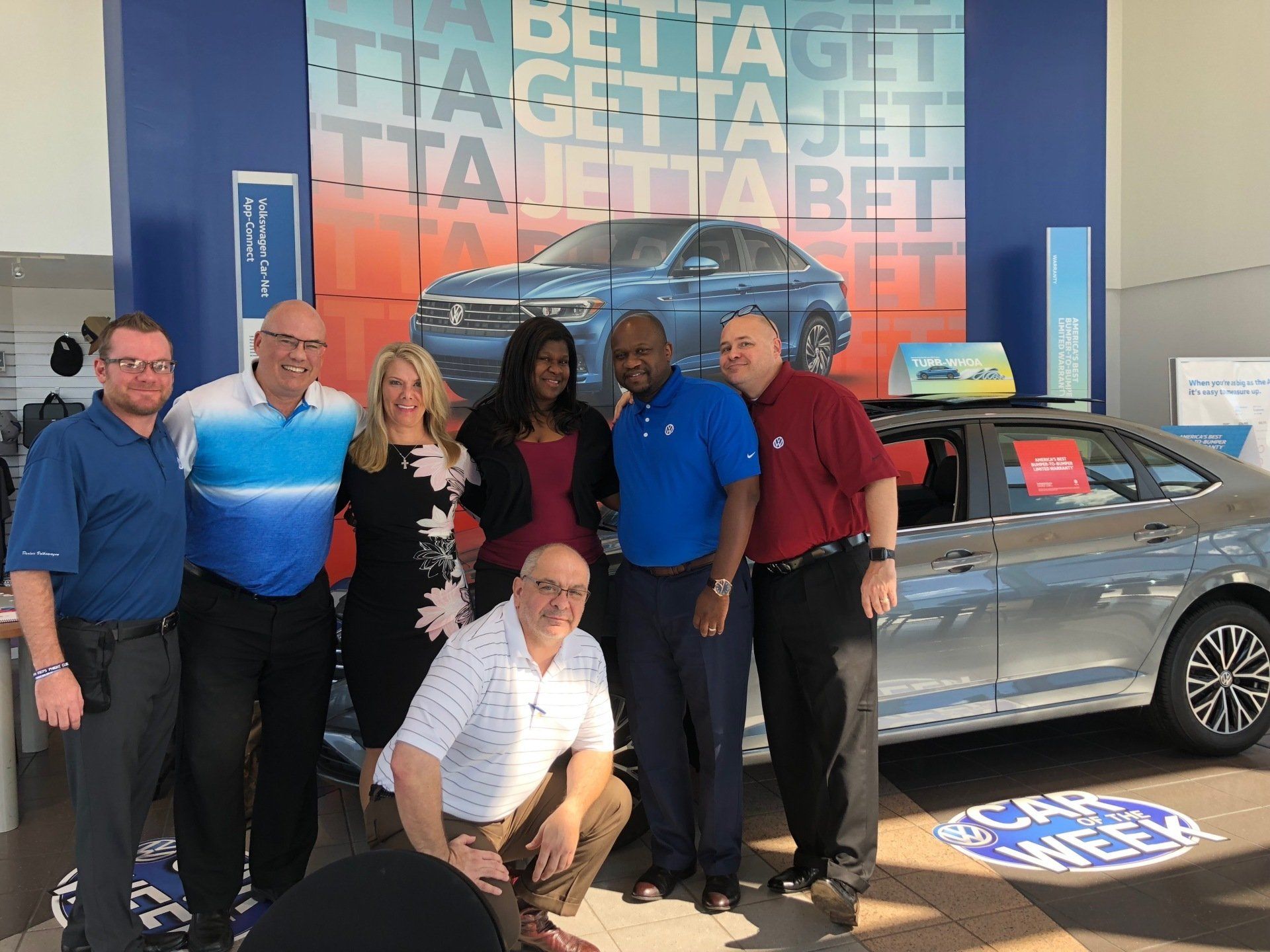Group of people posing near a car in a dealership. A blue and orange backdrop advertises 