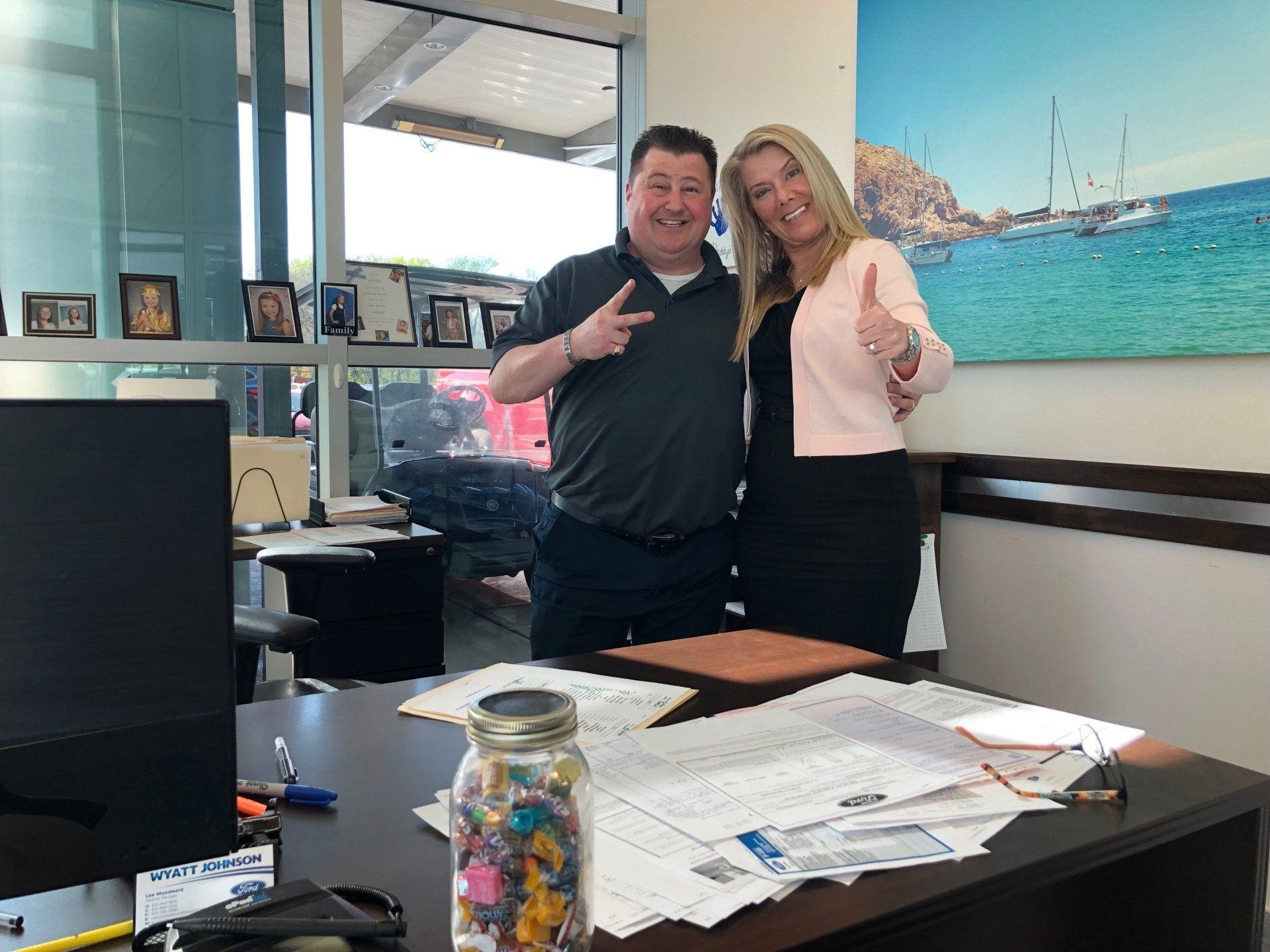 Two people smiling and posing in an office. One holds up a peace sign, the other a thumbs up.