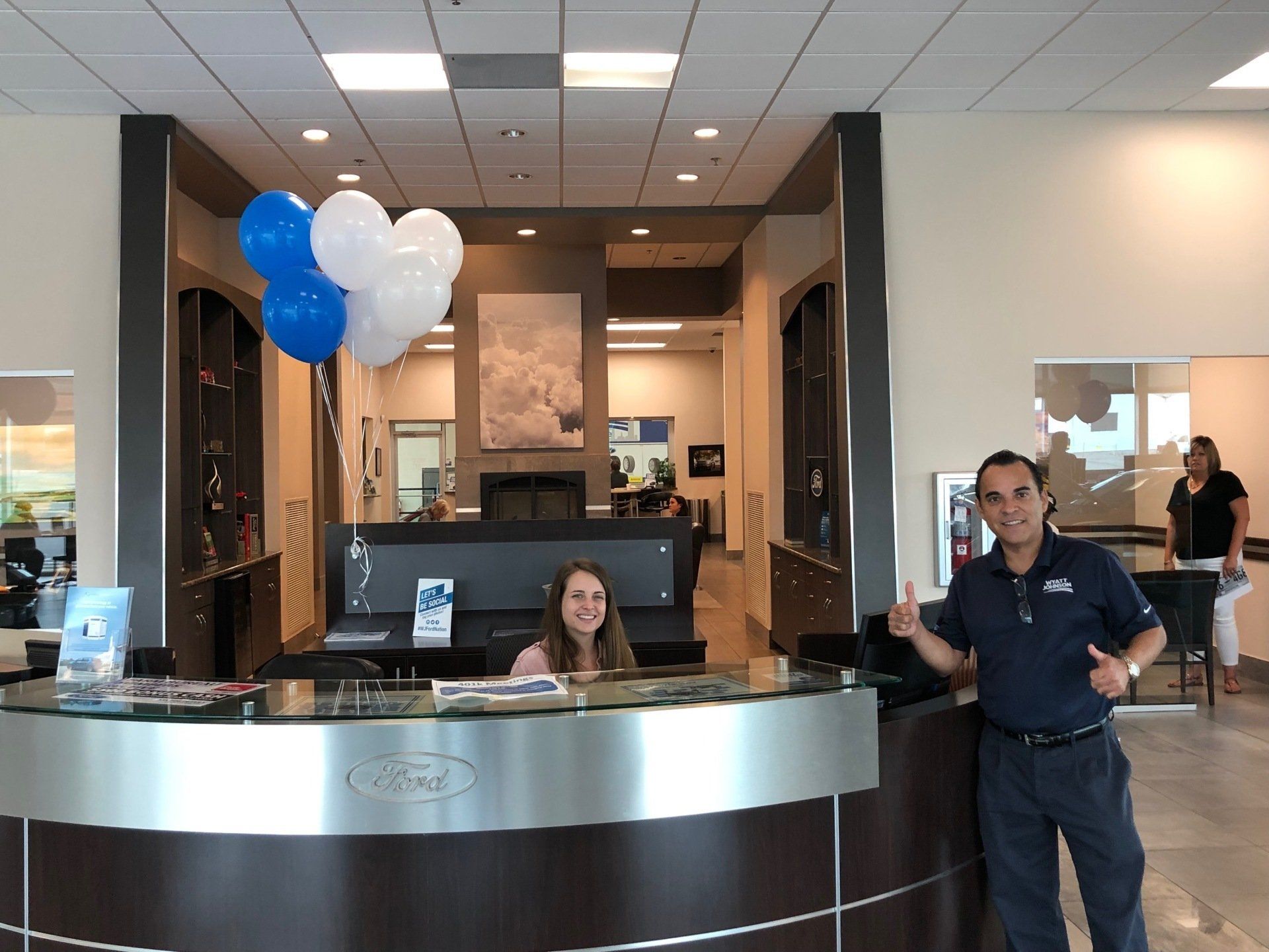 Two people at a reception desk; blue/white balloons, light interior, smiling faces.