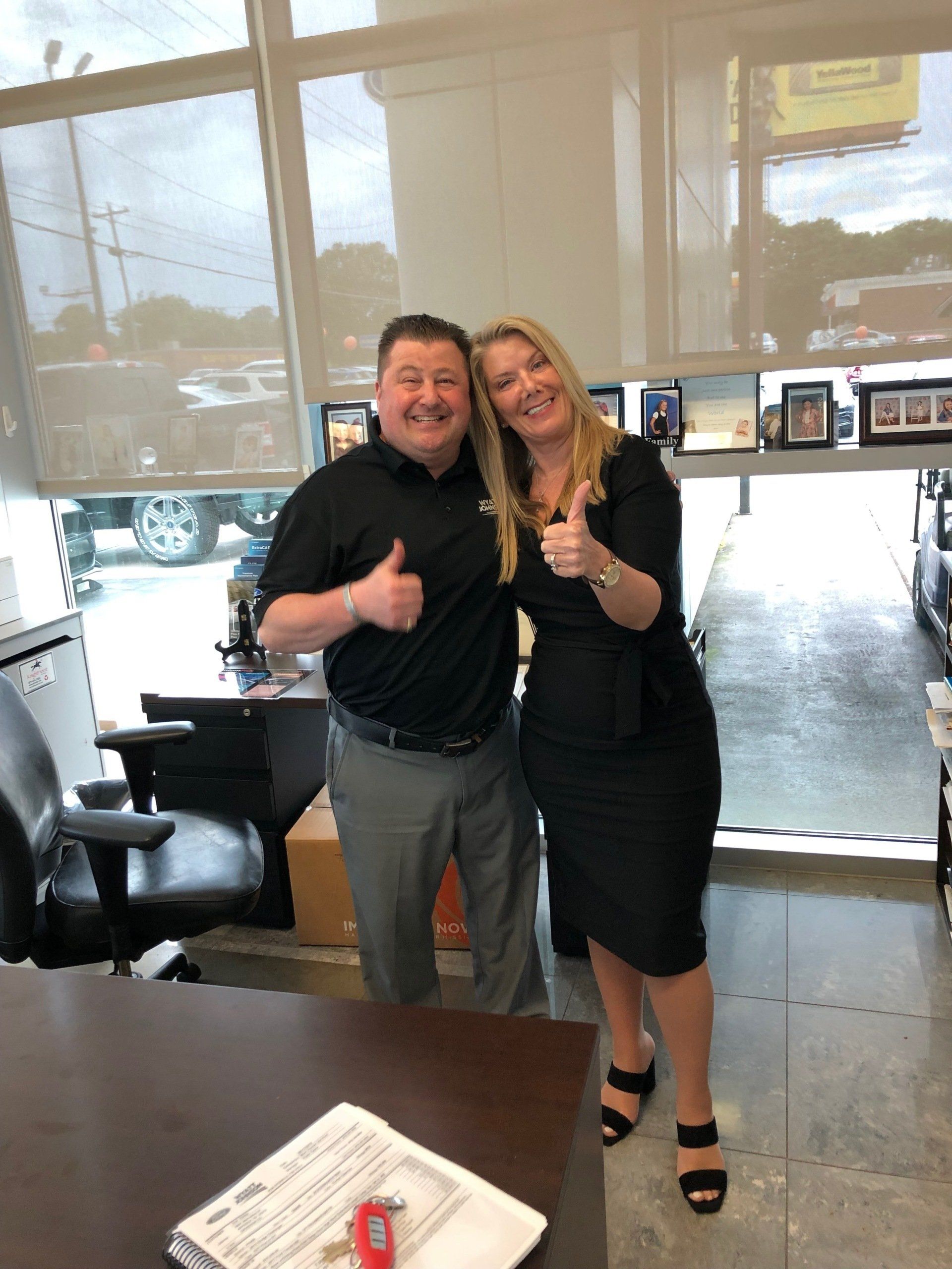 Man and woman giving thumbs-up, smiling. Inside a building, possibly an office, near a window.
