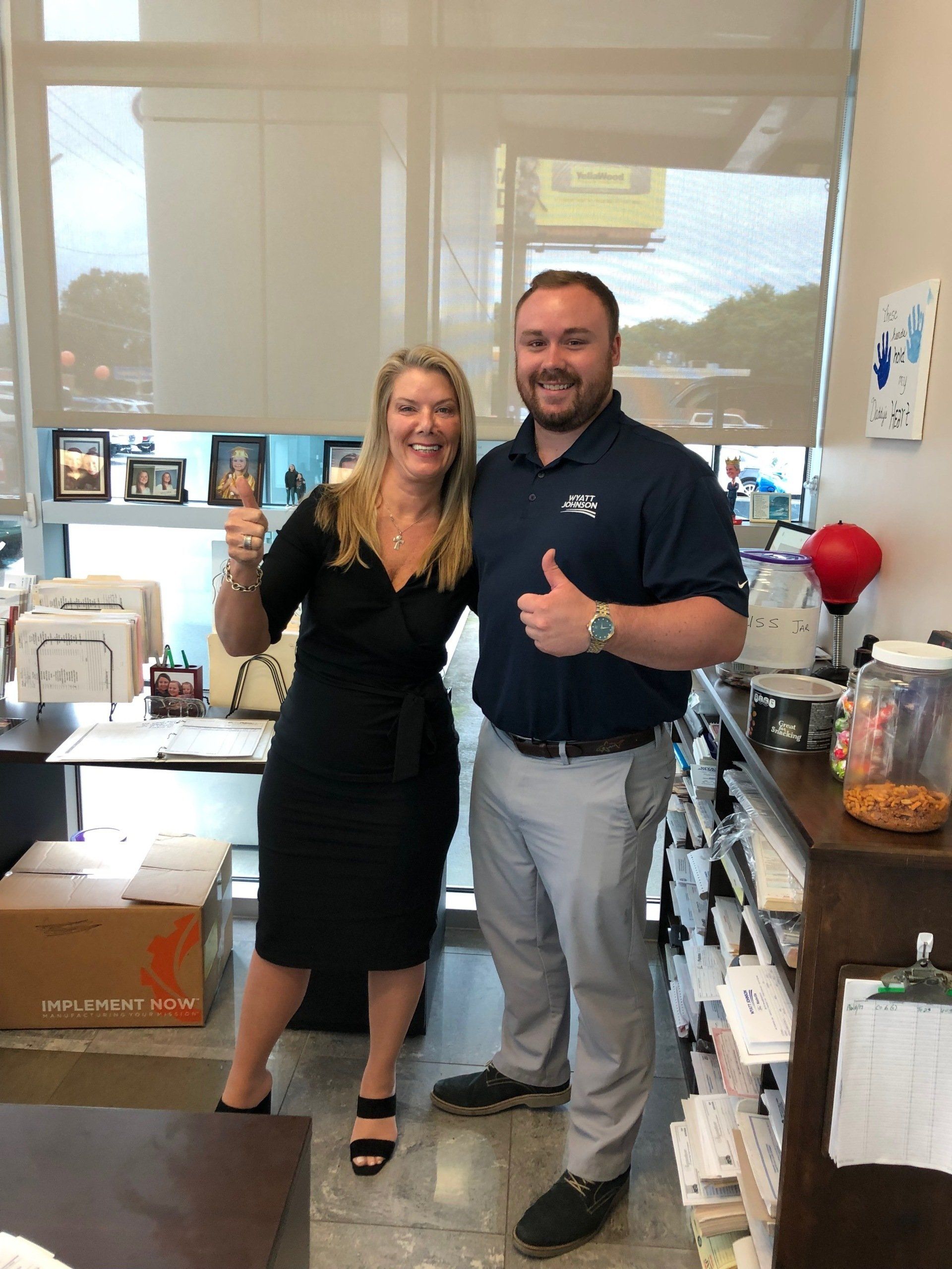 Two people giving thumbs up in an office. Woman in black dress, man in polo and grey pants.