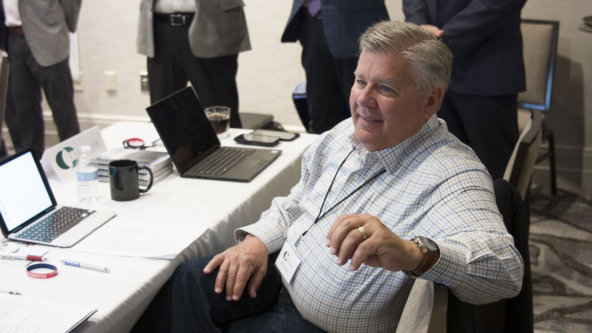 Man in a light-blue patterned shirt sits at a table with laptops, smiling. Other people stand behind him.
