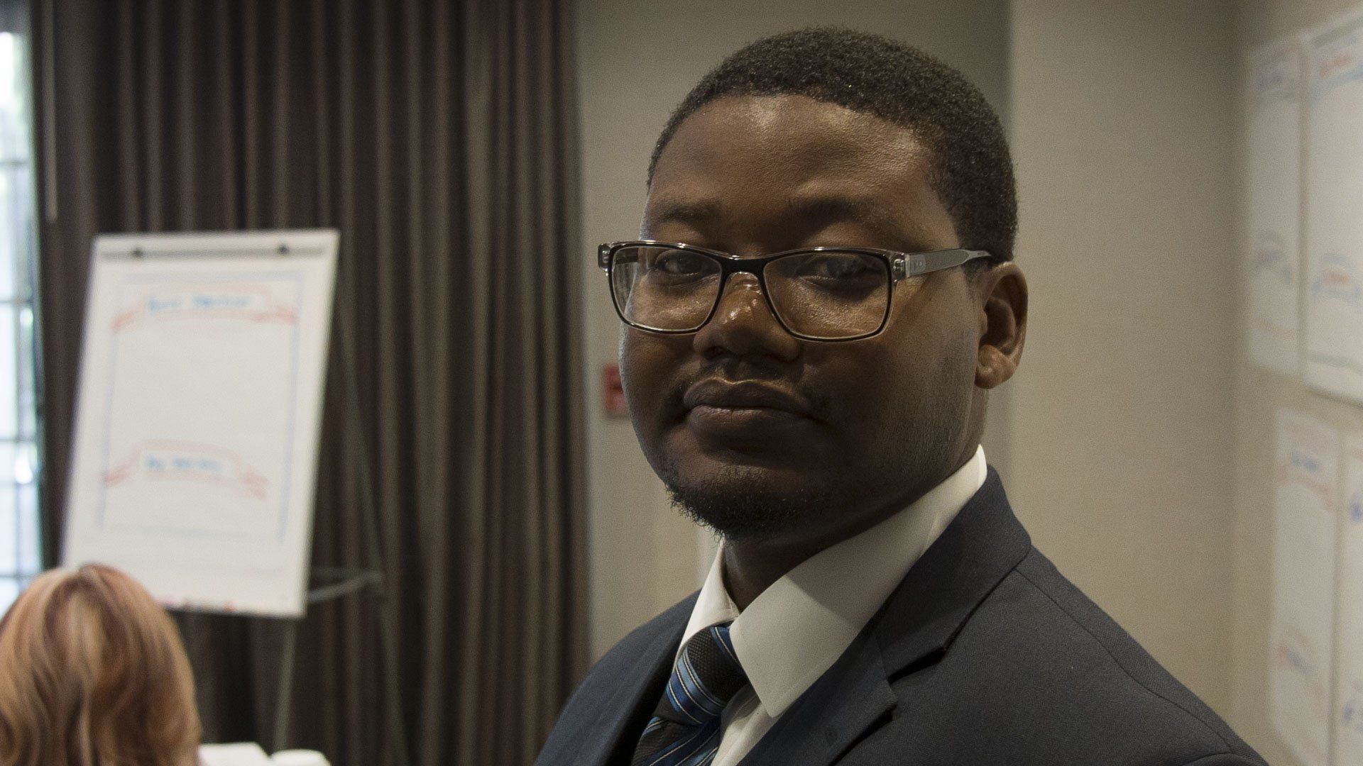 Man in suit and glasses, looking at the camera. He is in a meeting room with a whiteboard and curtains.