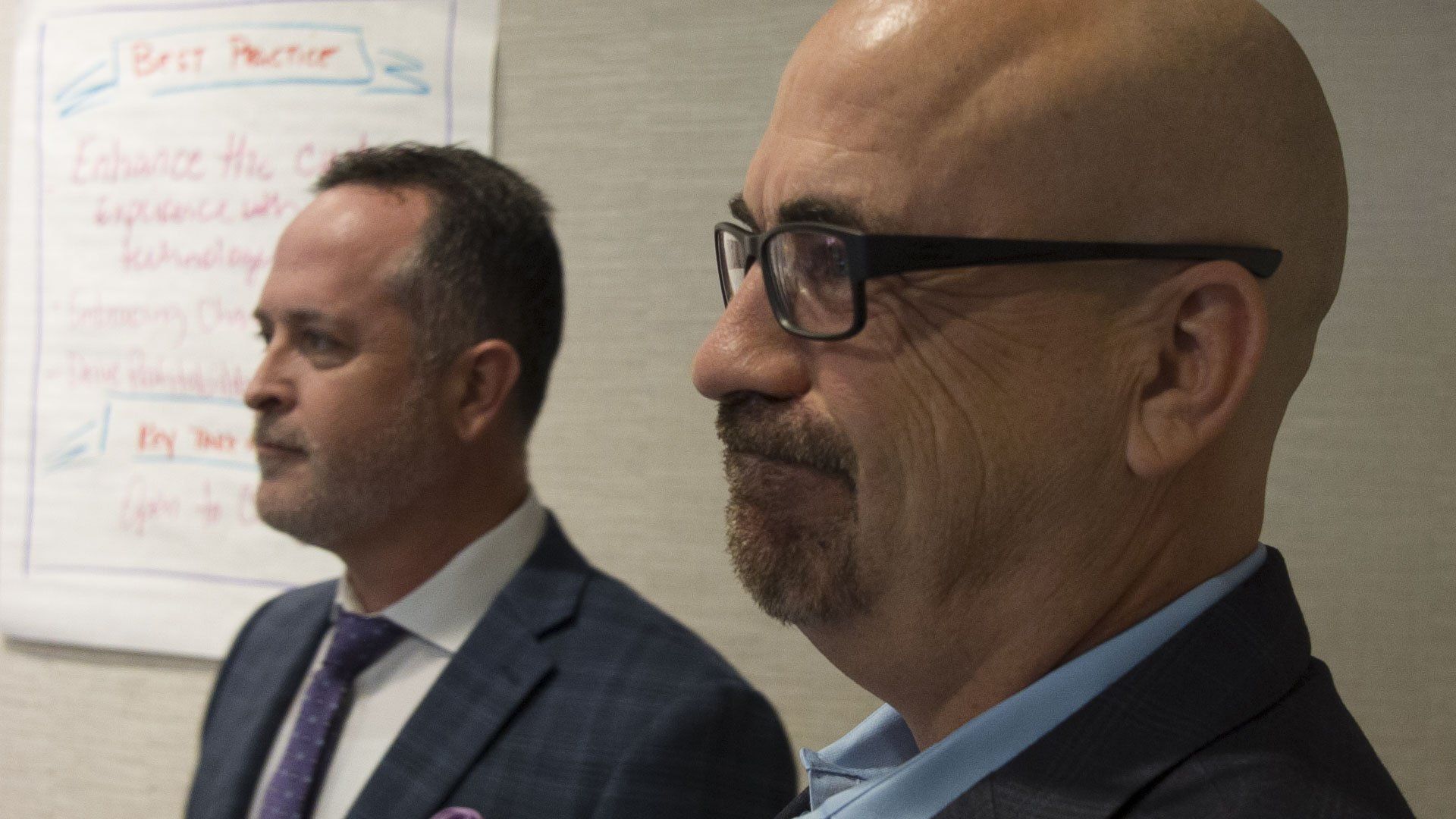 Two men in suits, one with glasses, looking to the side. A whiteboard is in the background.