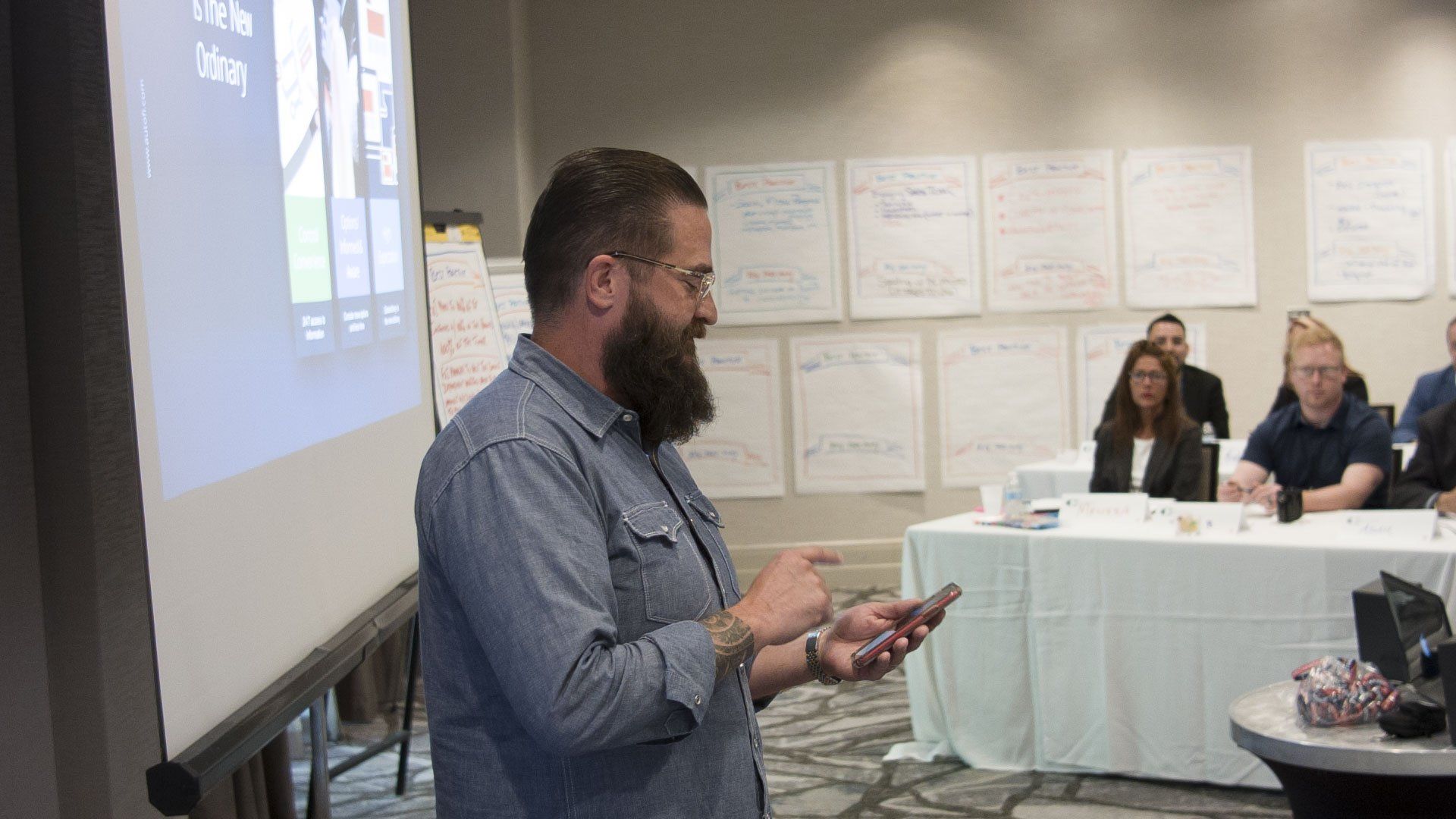 Man with beard presents with phone in hand, near a projector screen. People sit at tables, and papers are on the wall.