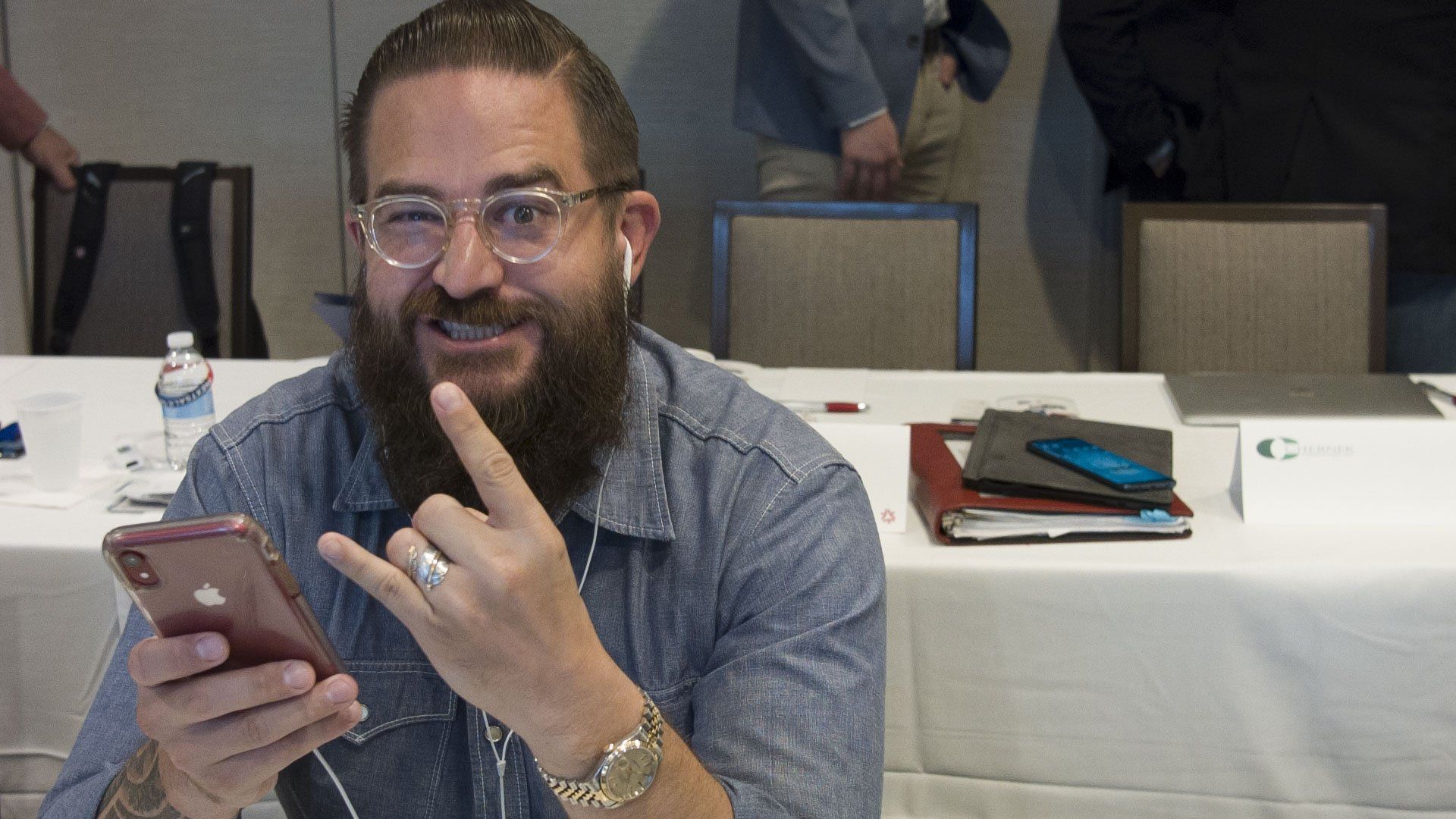Man with beard and glasses holding a phone, making a hand gesture, and smiling.