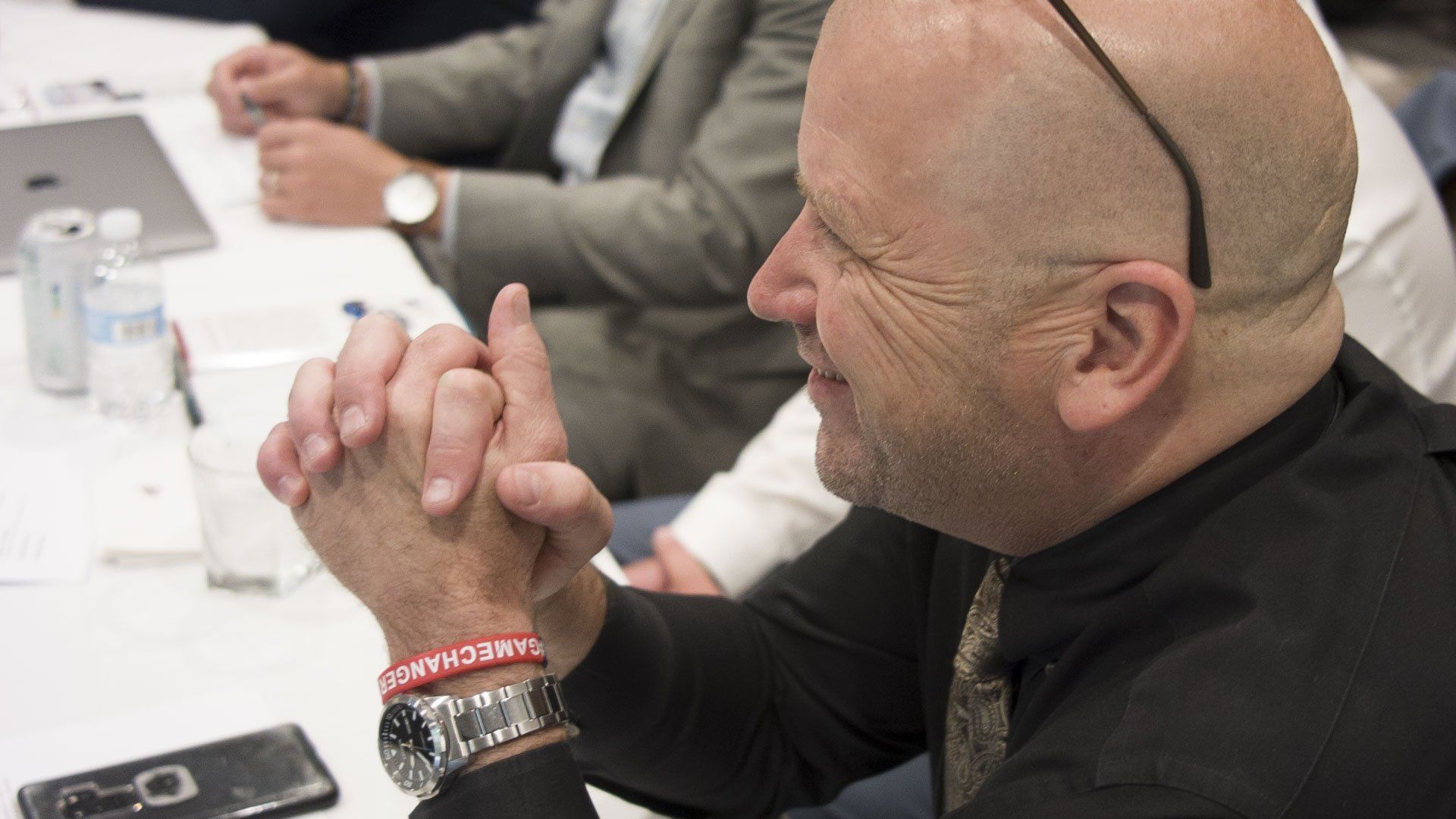 Bald man clasps hands in front of him, smiling. He wears a dark shirt and a watch.