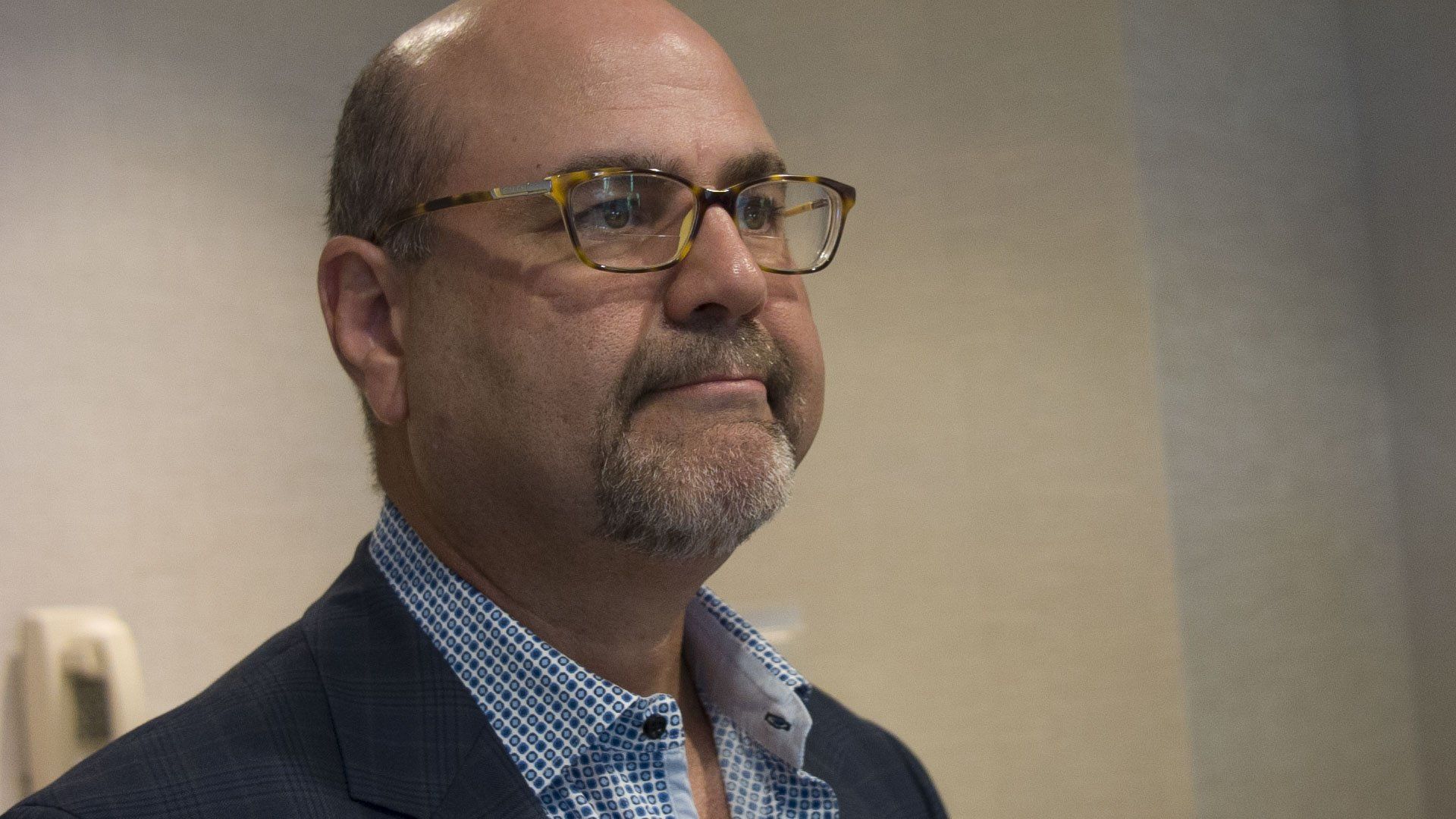 Man wearing glasses and a blazer looks slightly to the side in a room.