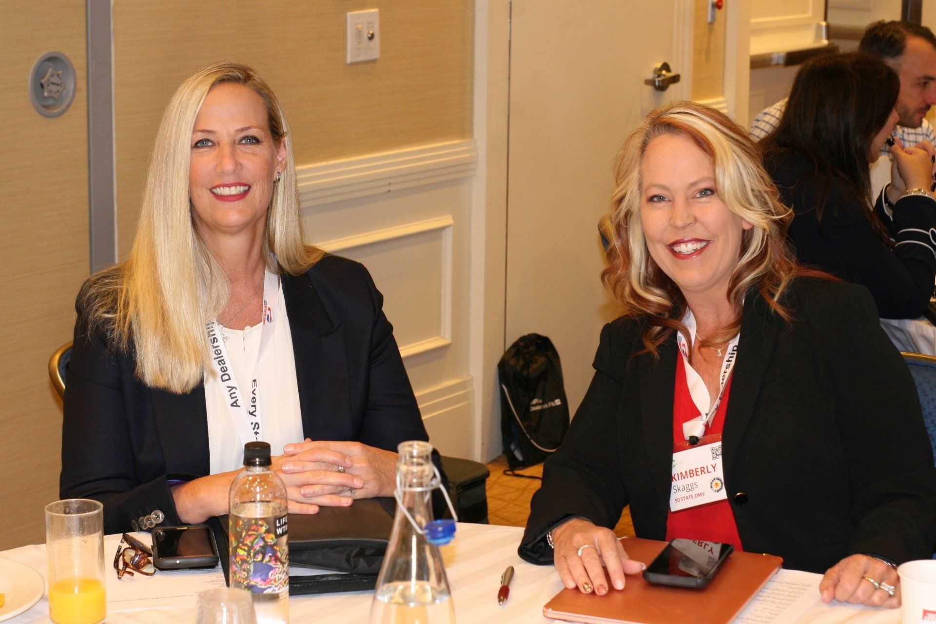 Two women seated at a table, smiling at the camera. They wear blazers, appear to be at a conference.