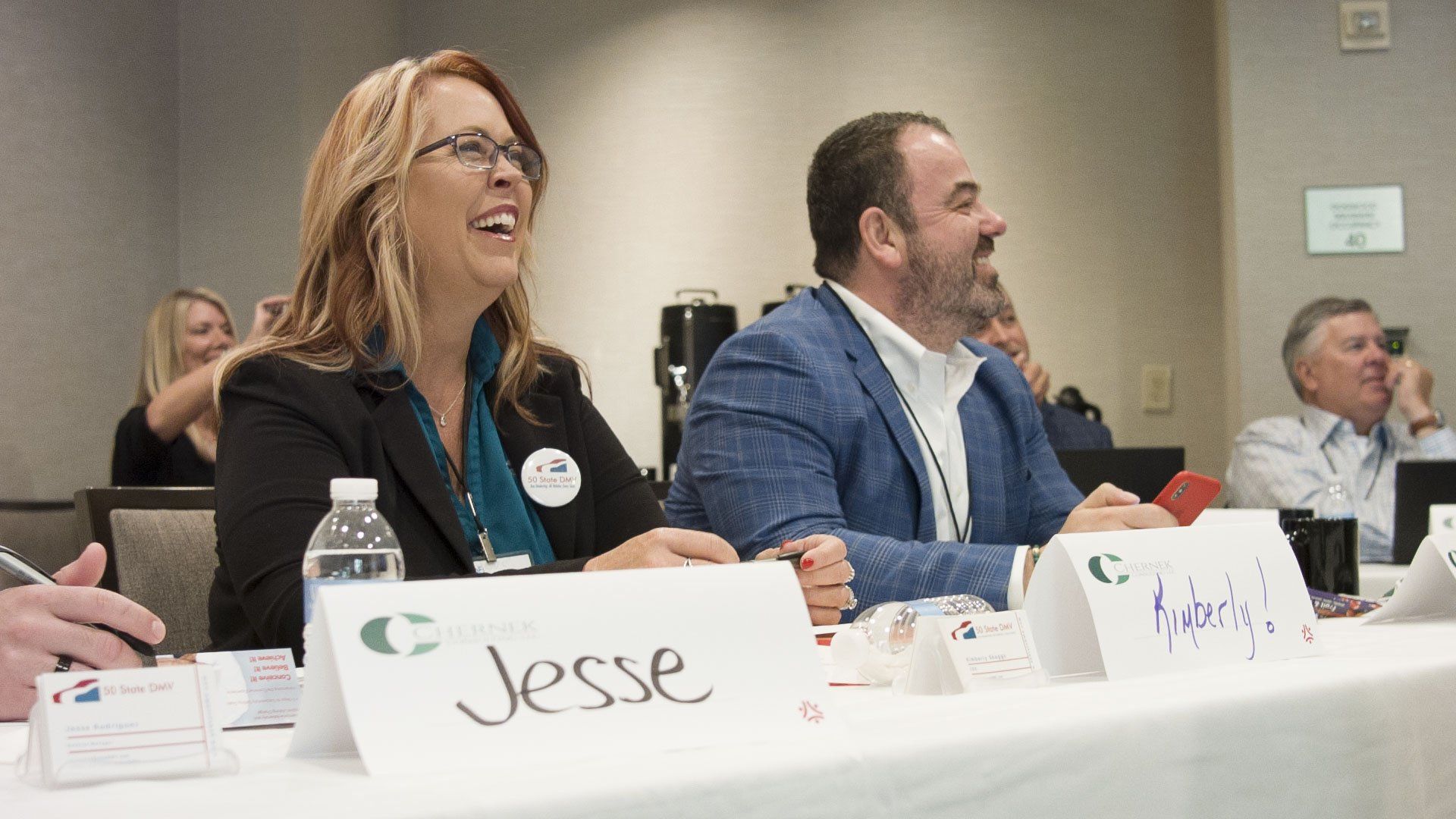 People at a conference table laughing; woman in black blazer, man in blue jacket, name tags visible.