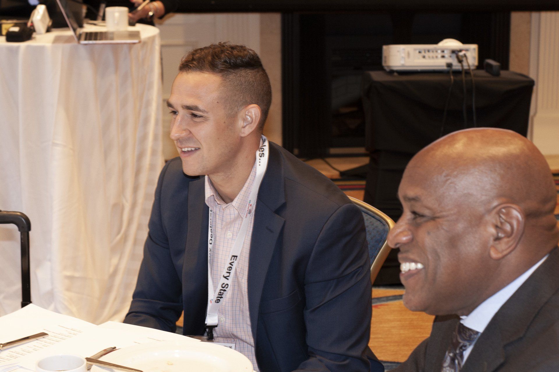 Two men at a conference table, smiling. One in a suit, the other in a blazer.