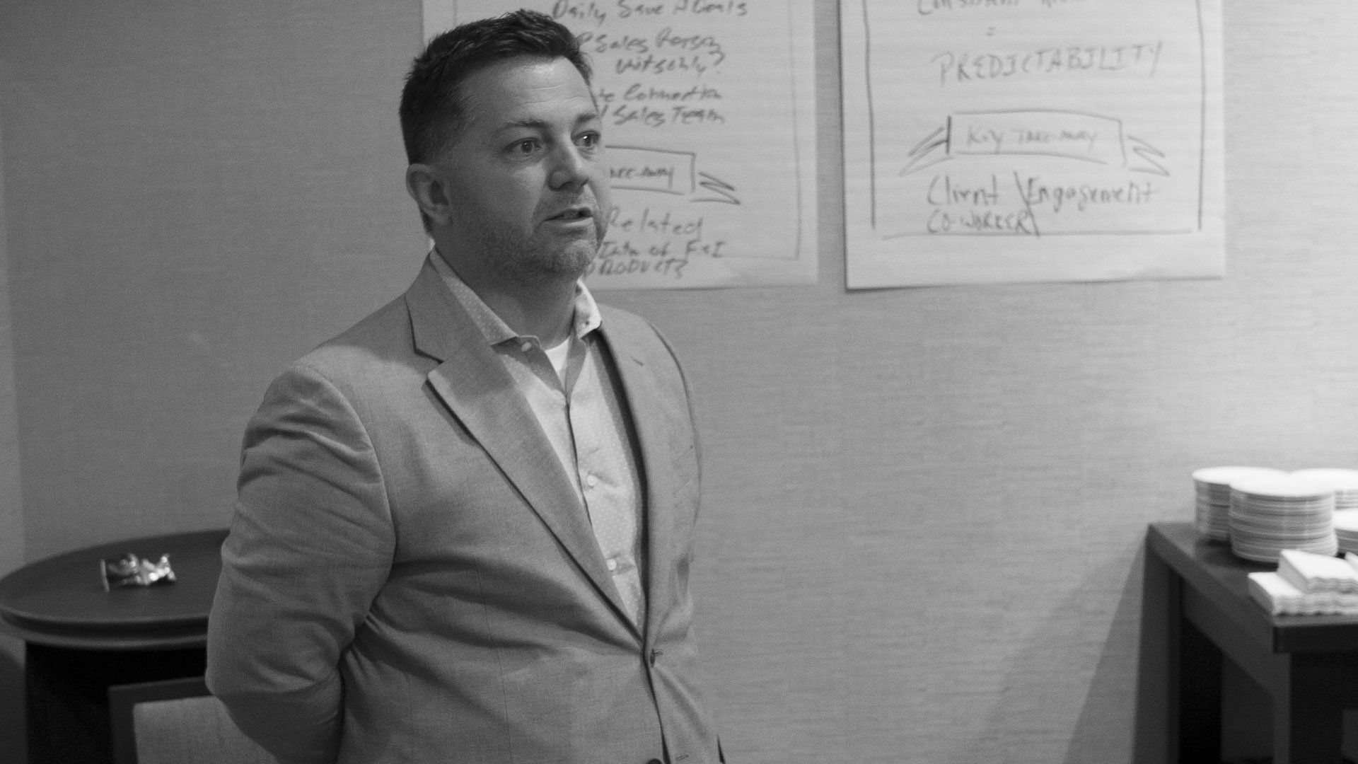 Man in a suit speaks, papers on wall behind him.