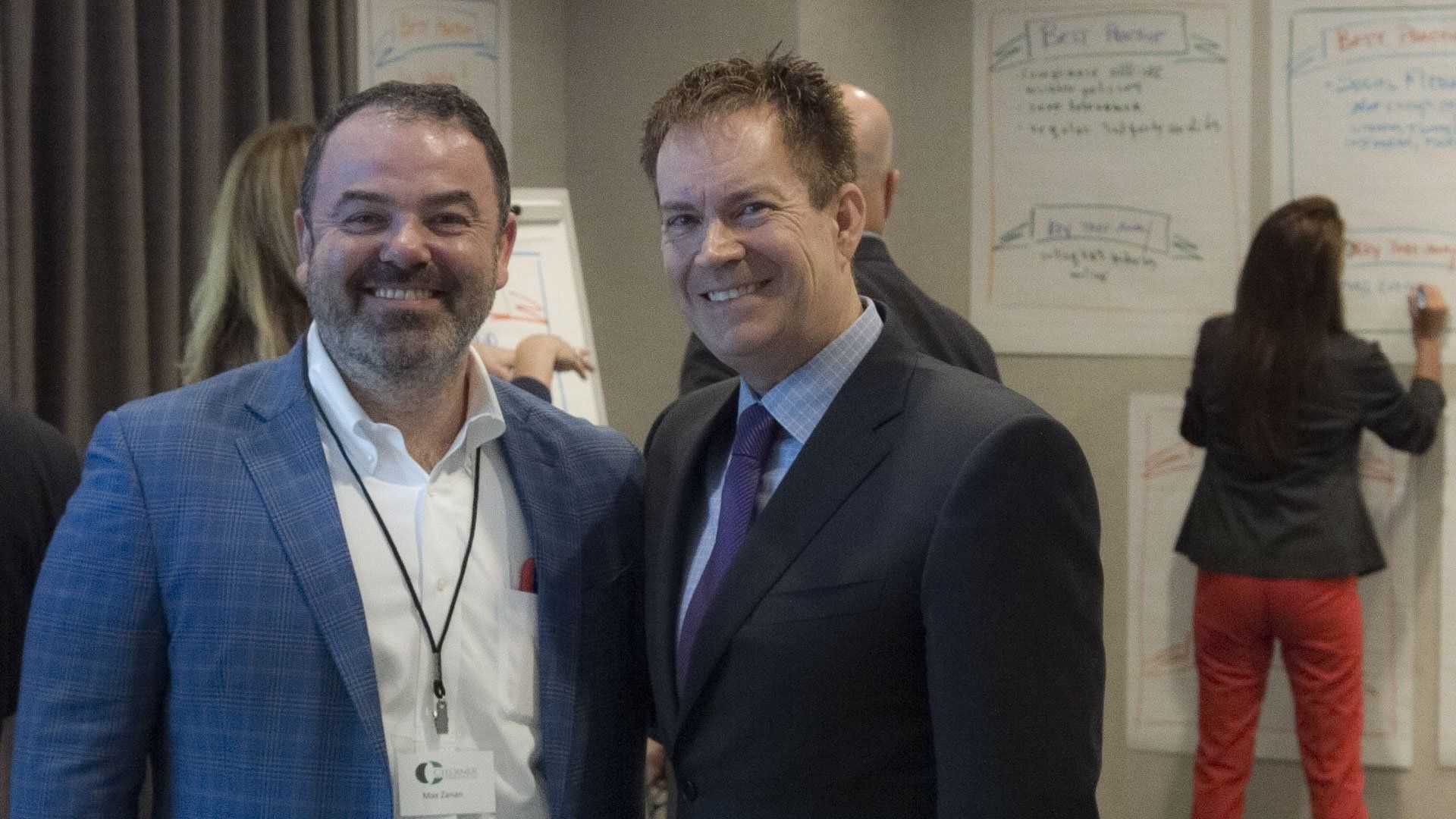 Two men smiling at a workshop, standing near flip charts with notes. Other people are in the background.