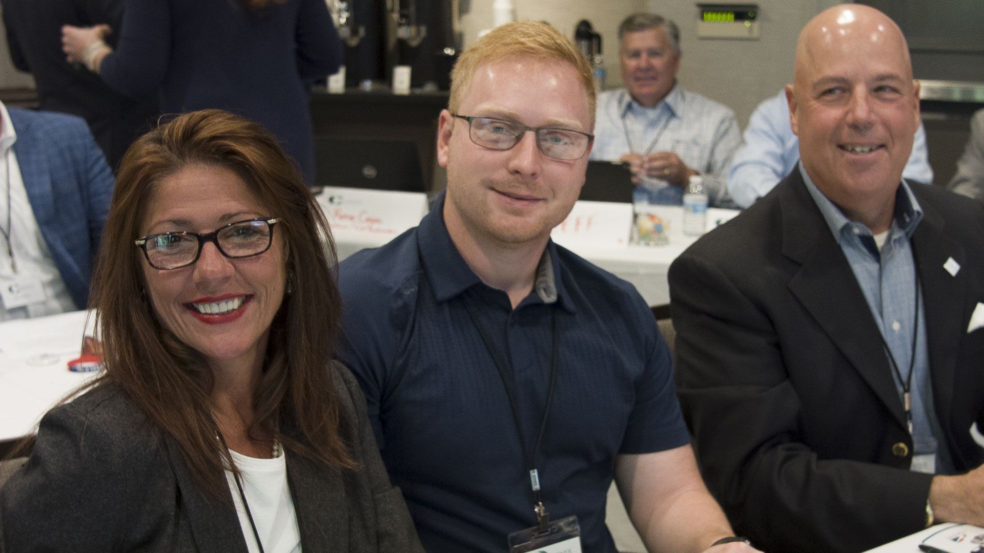 Three people smiling at a table, two wearing glasses. Others in background.