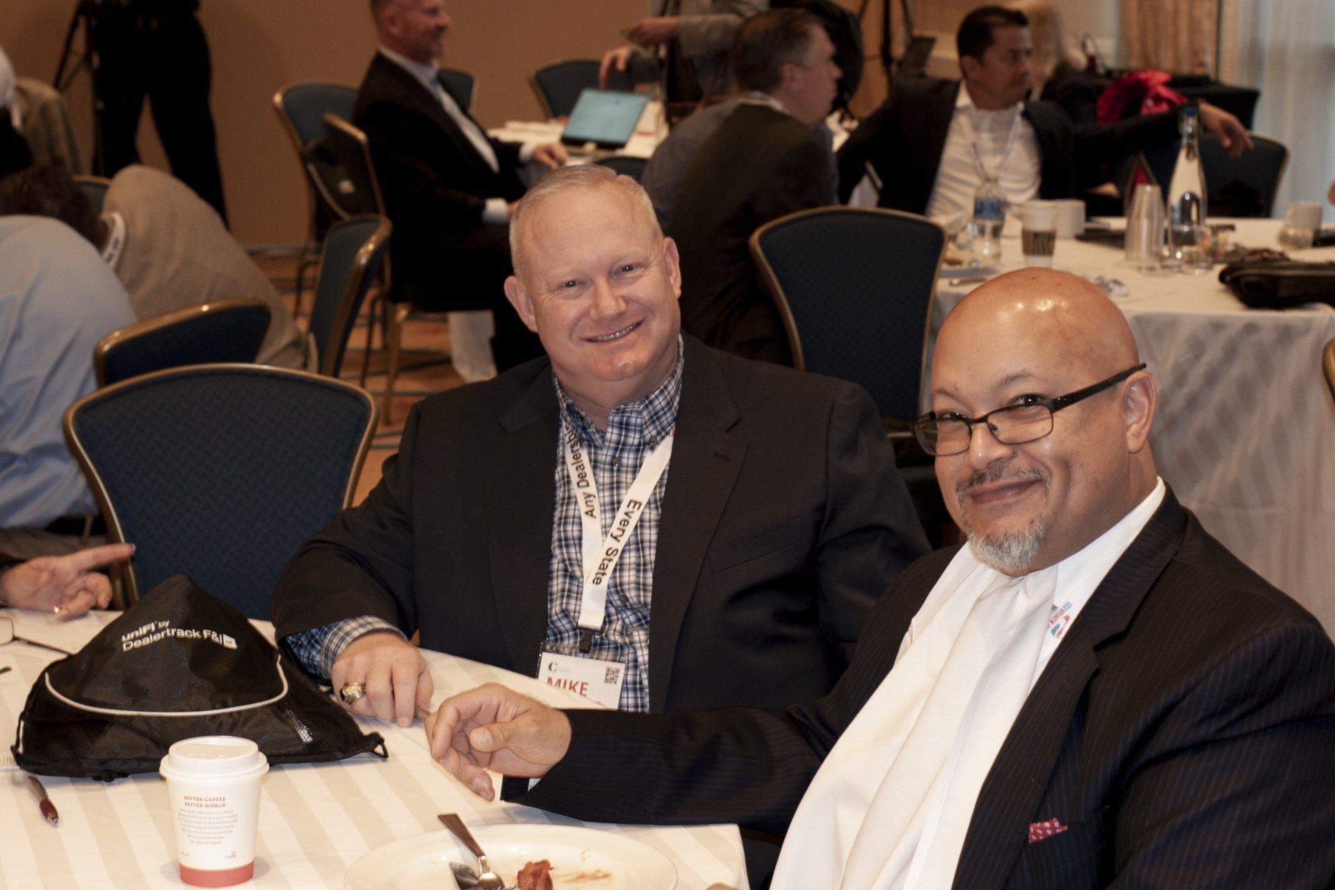 Two men at a table, smiling at the camera. They are in a conference room with others.