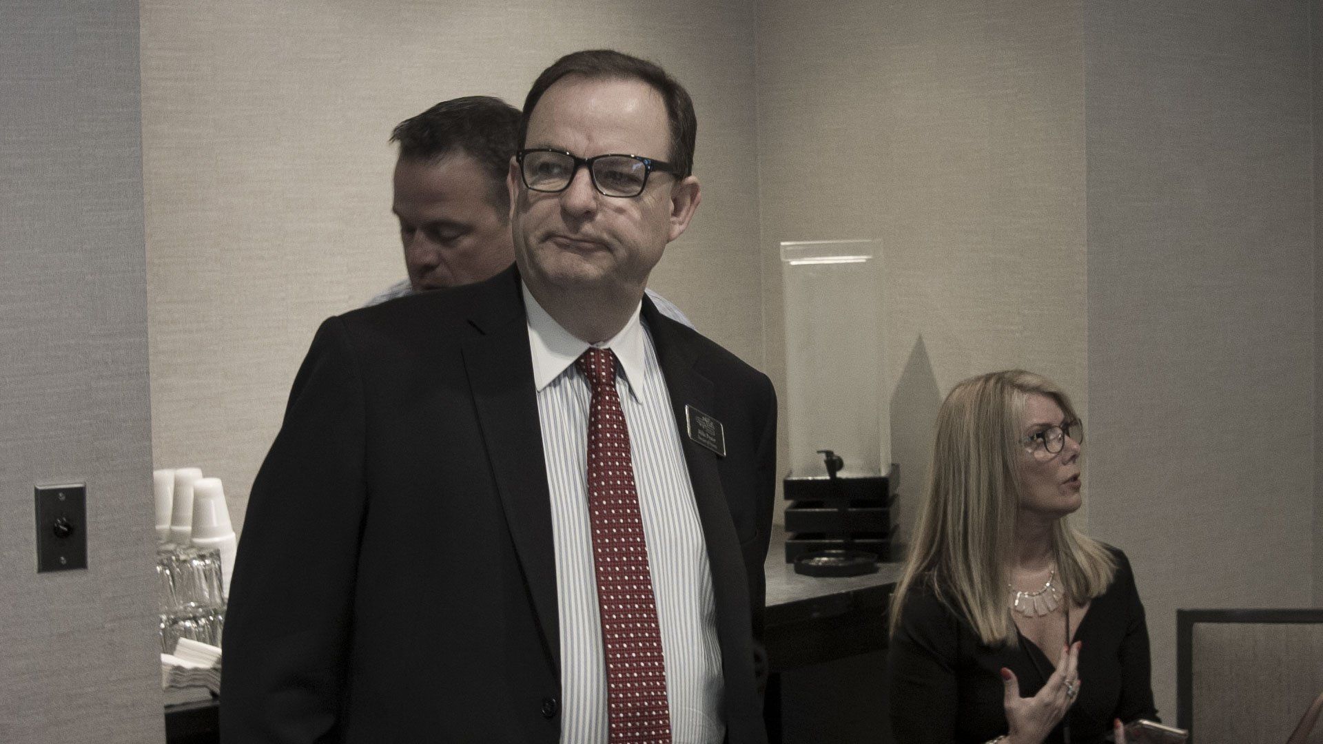 Man in suit and glasses looking annoyed, standing in a room with a woman talking.