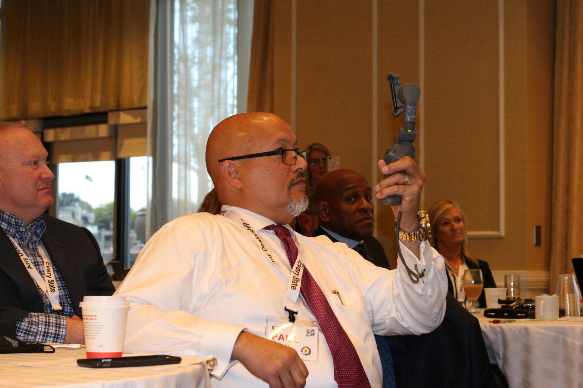 Man in white shirt holds up a device at a conference, others observe at a table in a bright room.