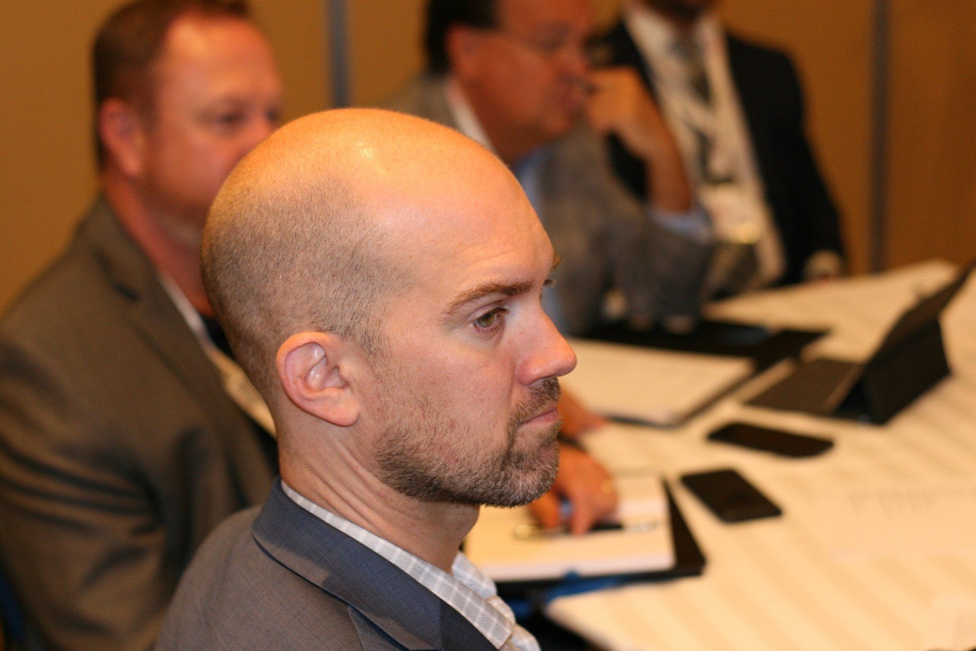 Man with shaved head at a meeting, looking to the side. Others sit at a table in the background.