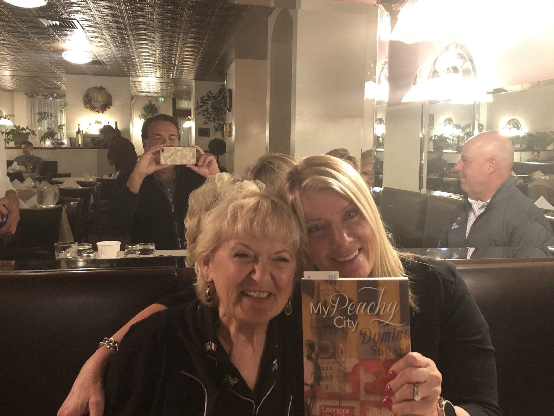 Two women smiling, hugging in a restaurant. One holds a menu, the other has her arm around her. Background: other diners.