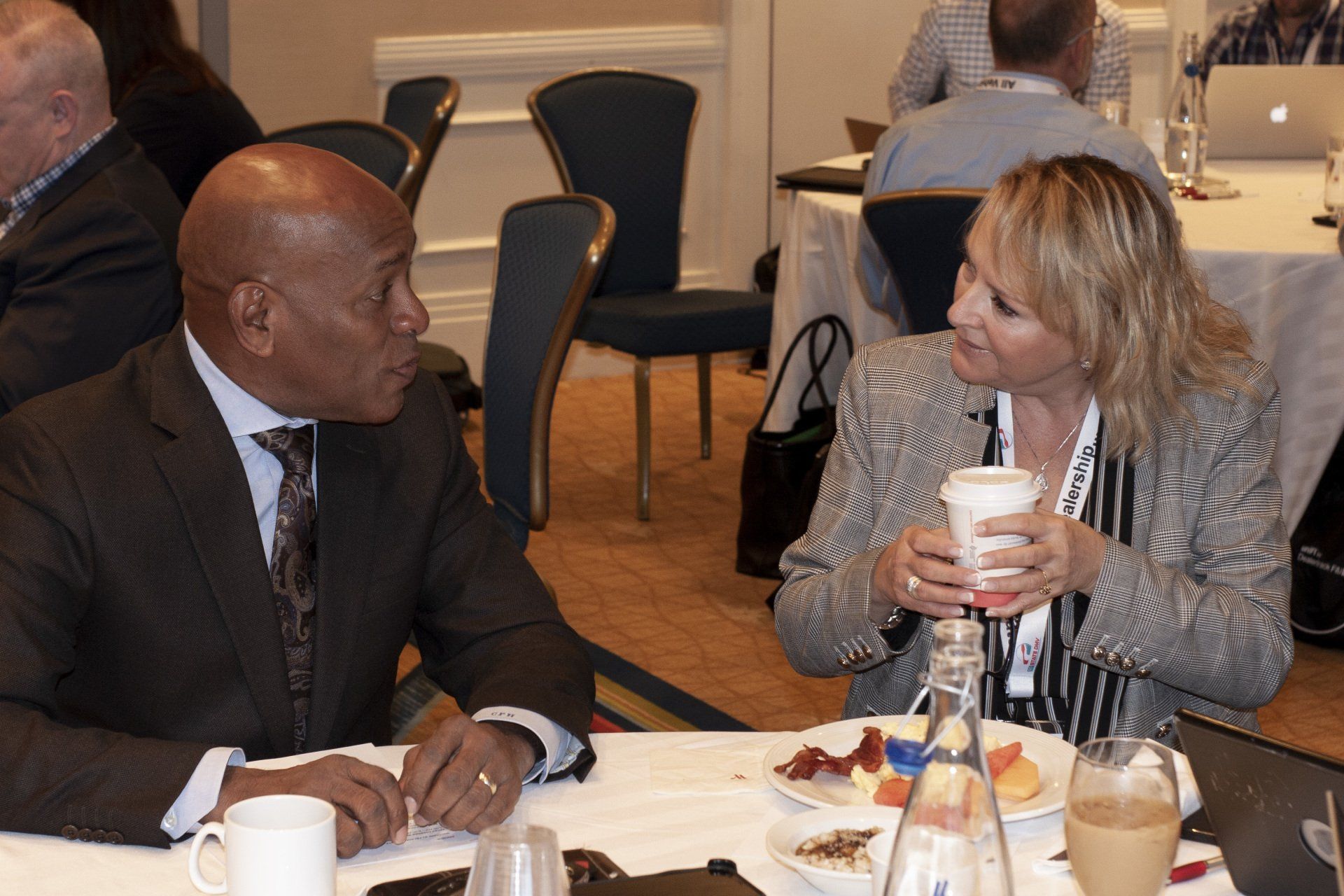 Man and woman seated at table, conversing. Woman holds coffee cup. Conference setting with others present.