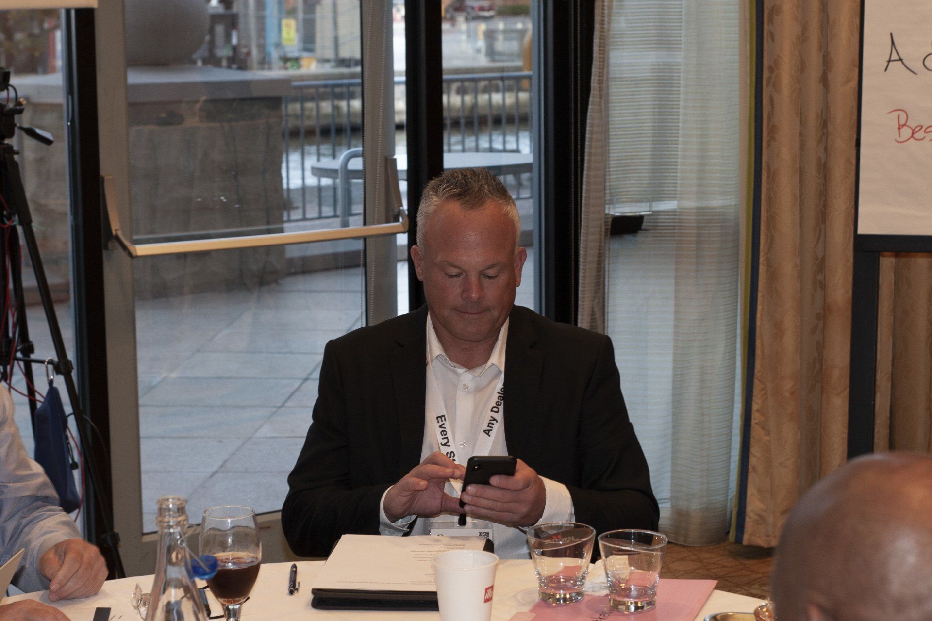 Man in black suit looking at a cell phone, seated at a table near a window.