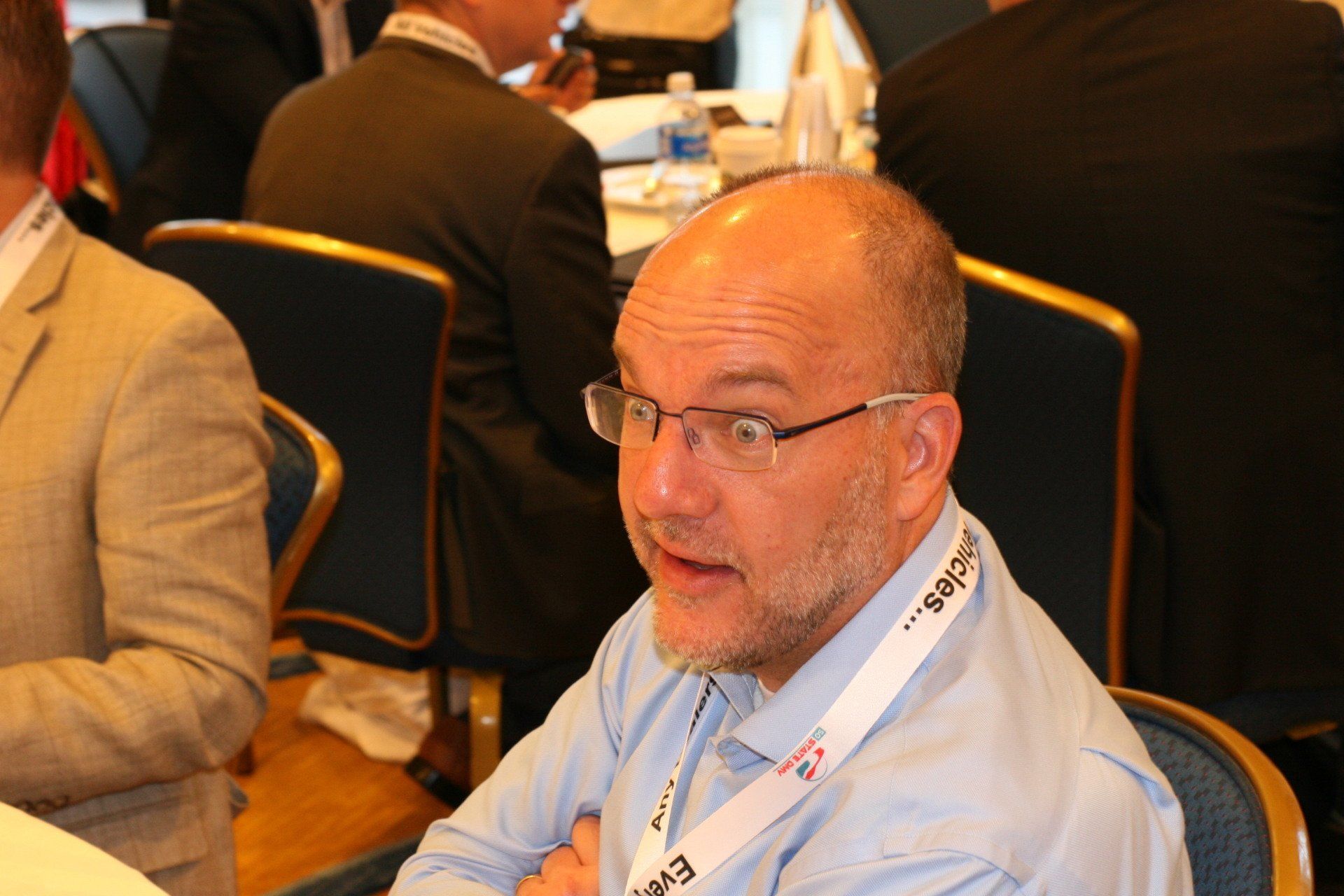 Man with glasses and a light blue shirt speaking at a conference, seated at a table.