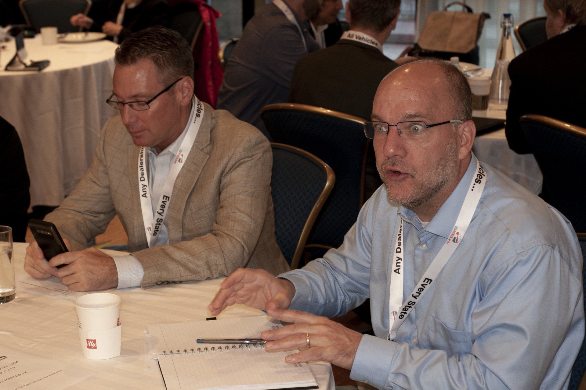 Two men in a conference setting. One uses a phone, the other gestures while speaking.