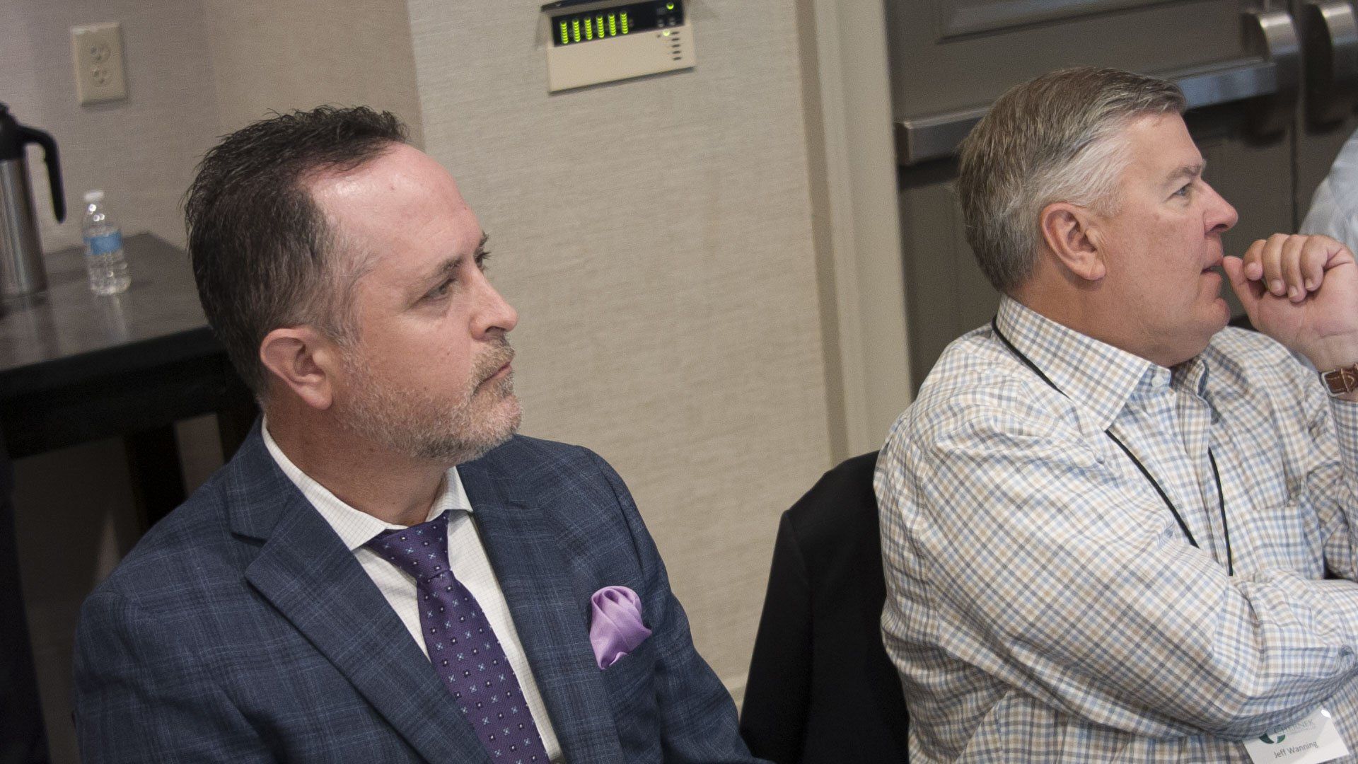 Two men attentively listen in a room. One wears a blue suit, the other a button-down shirt.