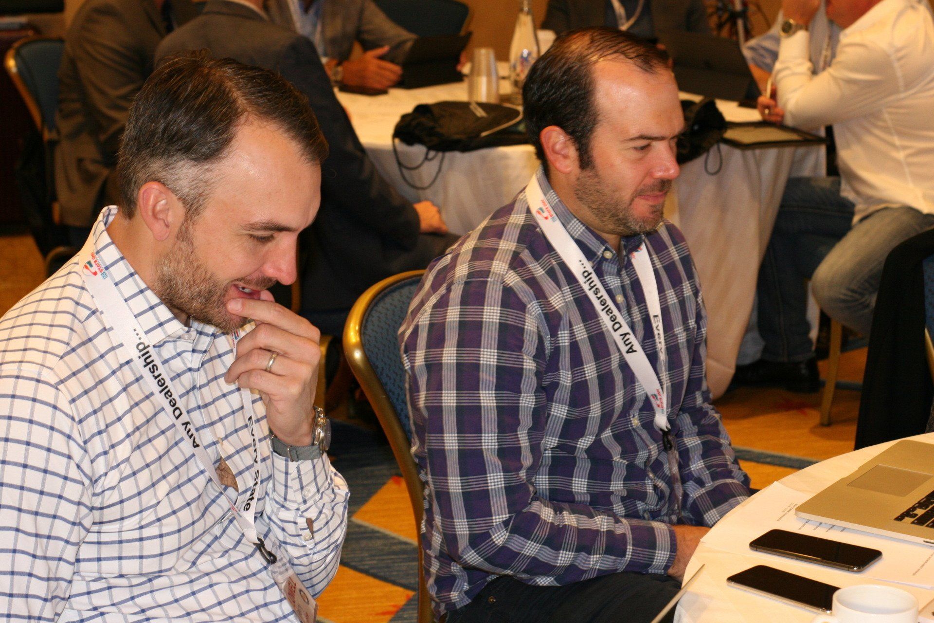 Two men at a table, looking down at objects. One bites a finger, the other wears a plaid shirt. Conference setting.