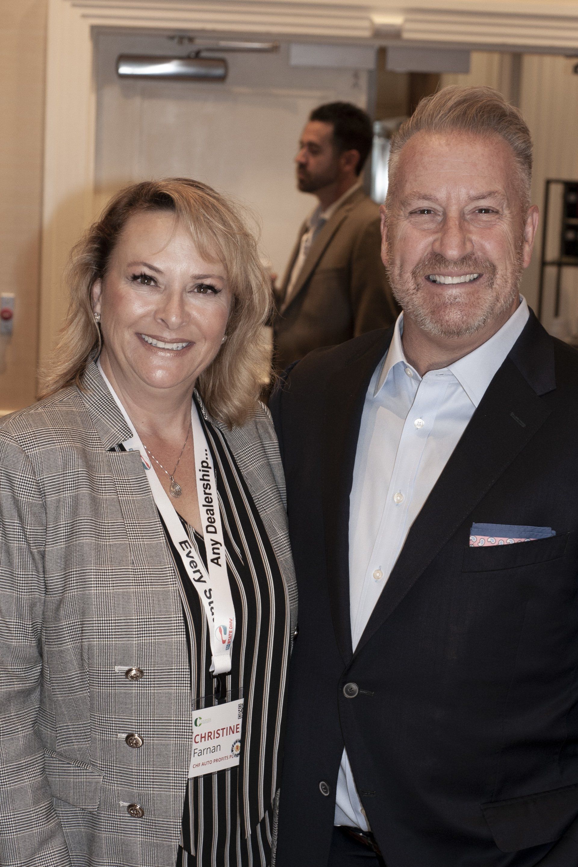 Woman and man smiling, standing close. She wears a black/white patterned jacket; he a dark suit.
