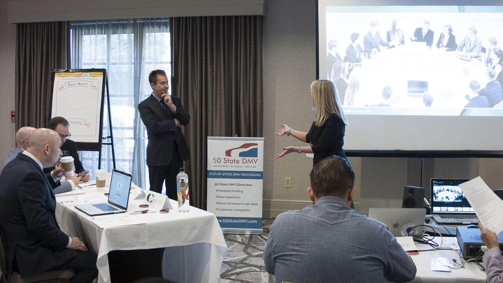 Business meeting with presenters, attendees around tables, projection screen, whiteboard, banners, and laptops.